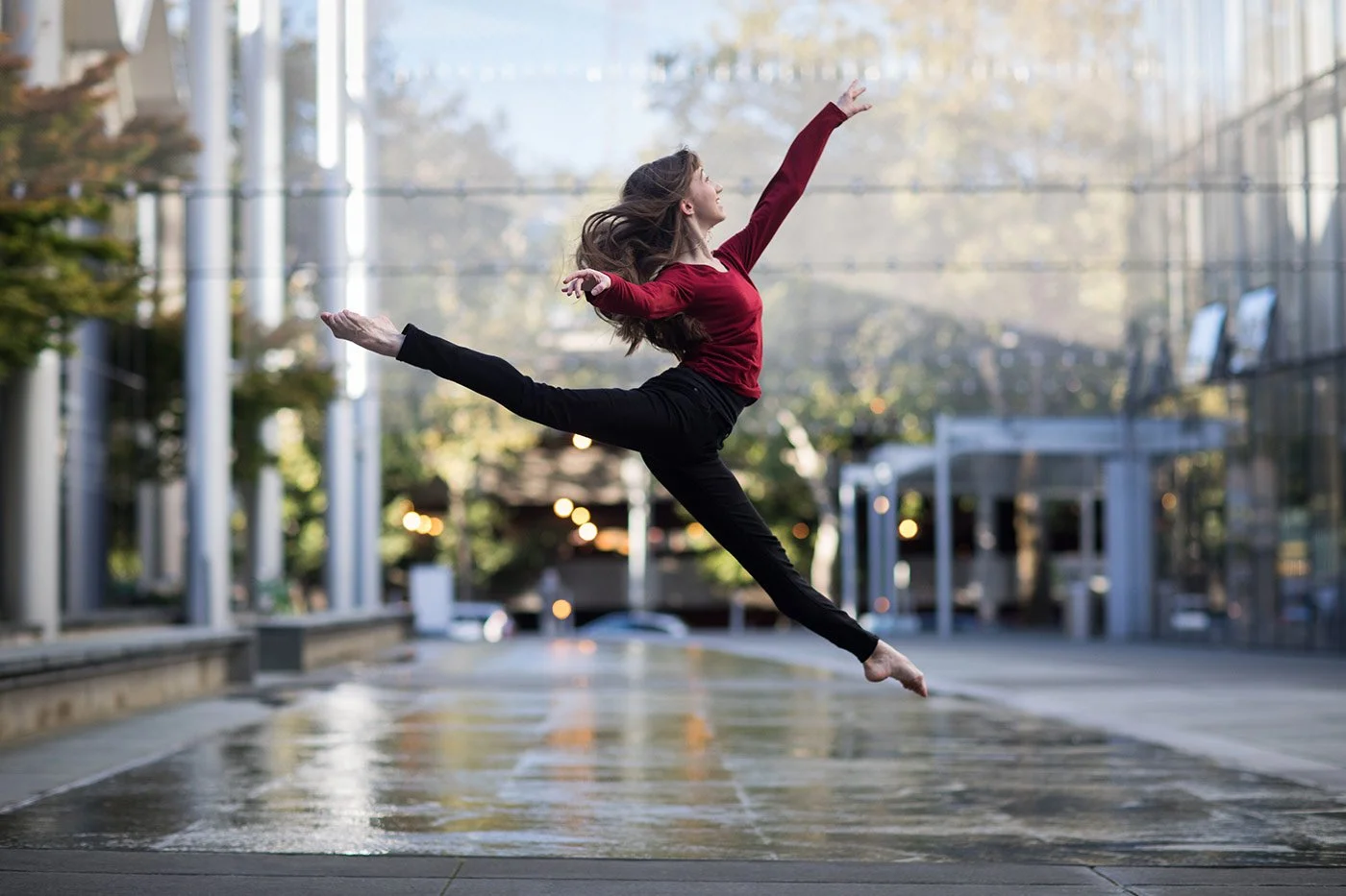 A woman dancing outdoors on a wet pavement in an urban area with modern glass buildings and trees, captured in mid-air leap.
