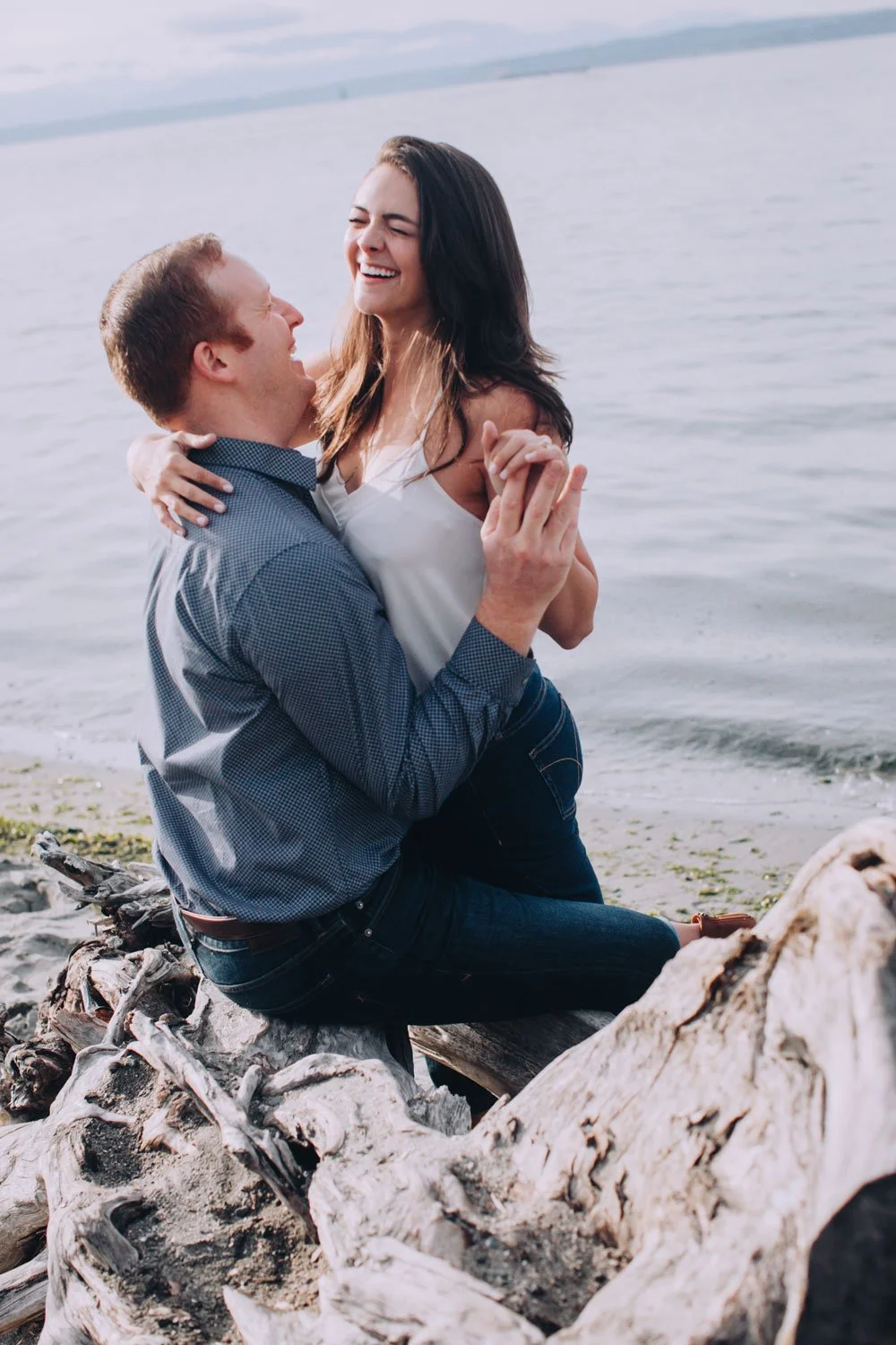 A man and woman sharing a joyful moment on a beach, with the man kneeling on driftwood and the woman smiling with her eyes closed, holding his hands, during daytime.