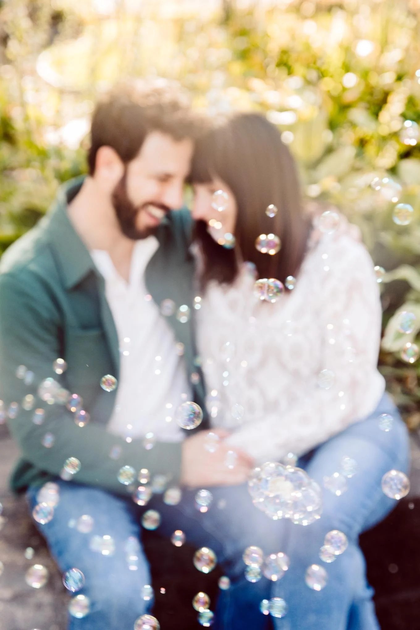 A couple sitting outdoors surrounded by bubbles, smiling with their heads together.