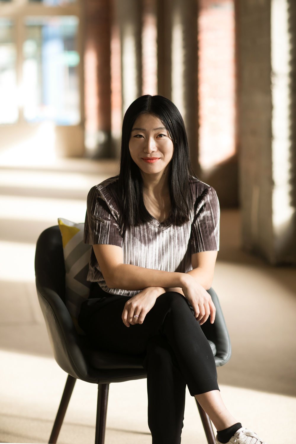 A young woman with long black hair, sitting on a black chair in a well-lit indoor space with large windows and brick walls, smiling and looking at the camera.