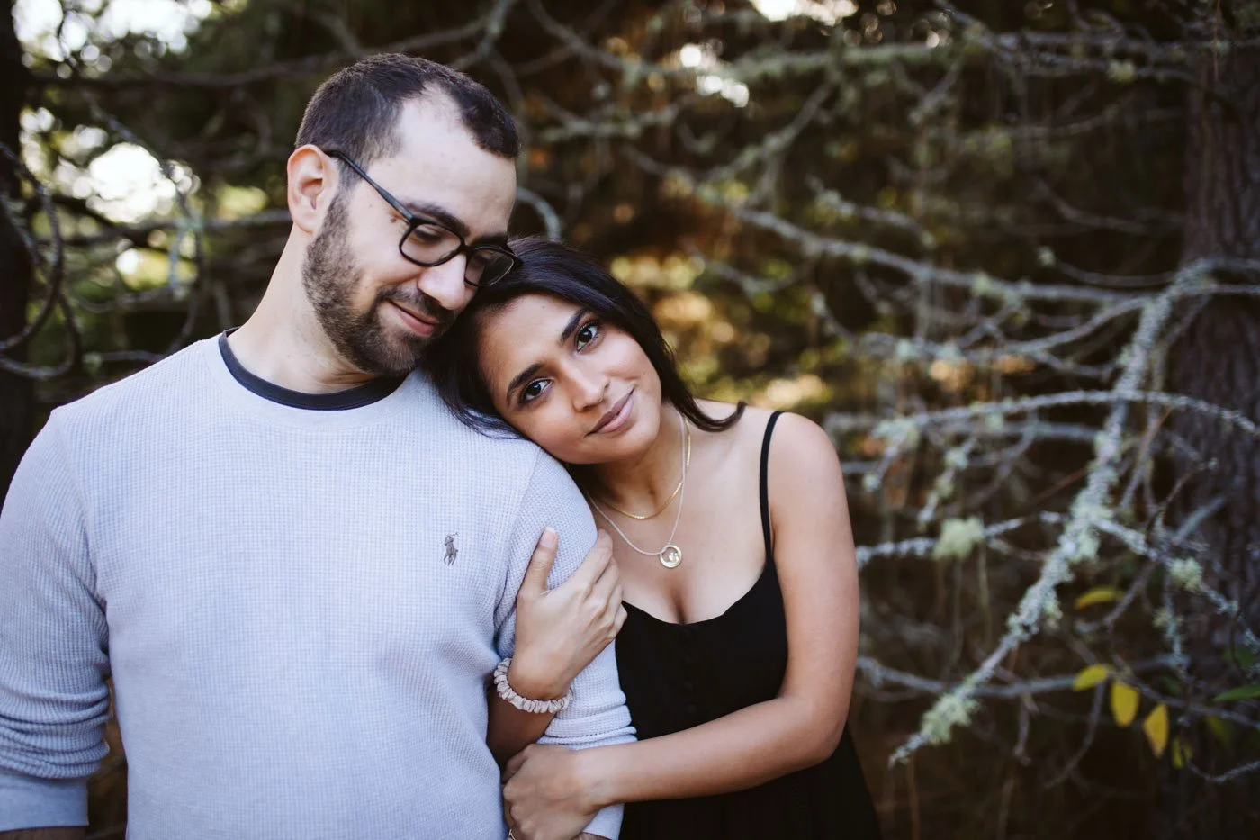 A couple at a wooded outdoor location, the woman with her head resting on the man's shoulder, both looking relaxed and content.