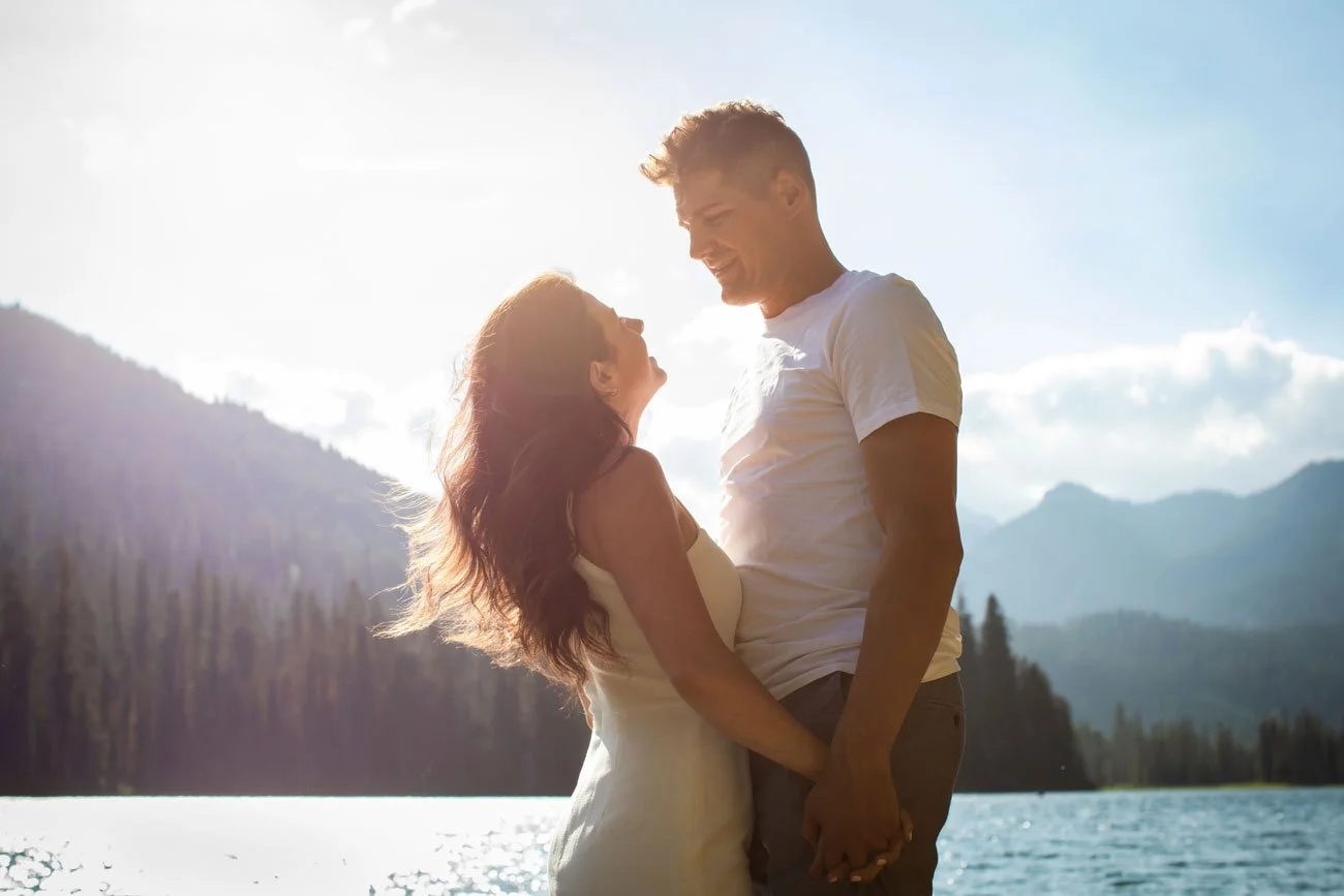 A couple standing close to each other outdoors, smiling and holding hands, with mountains and a lake in the background under a sunny sky.