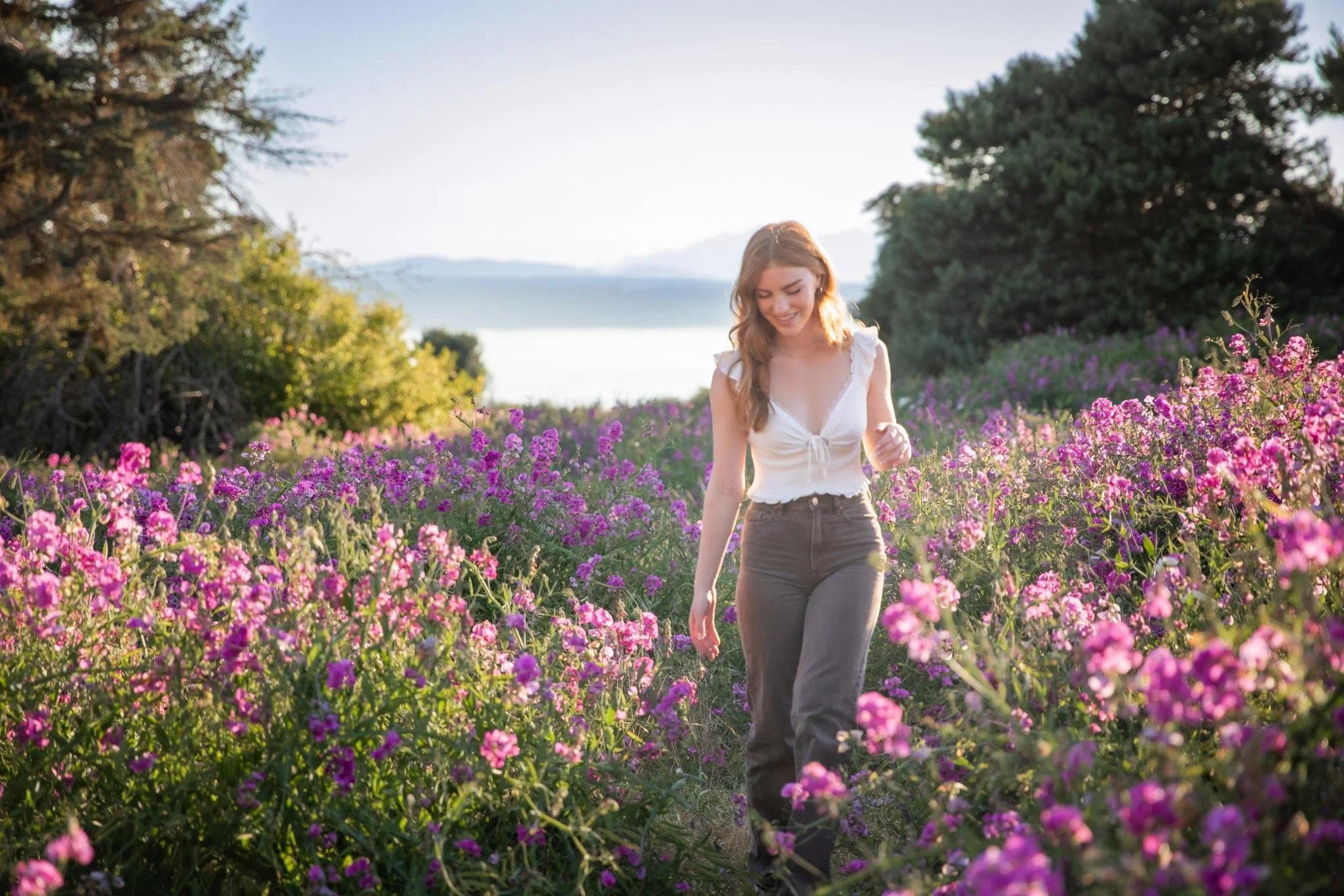 A high school senior with long wavy hair, smiling, walking through a field of pink and purple flowers, with trees and distant water in the background, during sunlight.