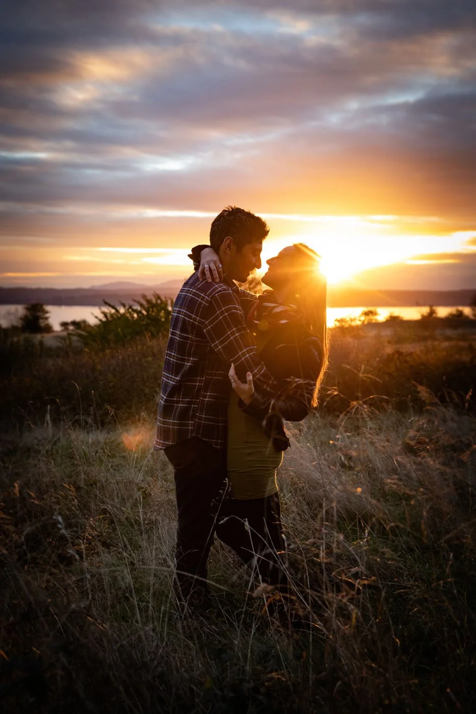 A couple embracing outdoors at sunset on a grassy field with water and distant mountains in the background.