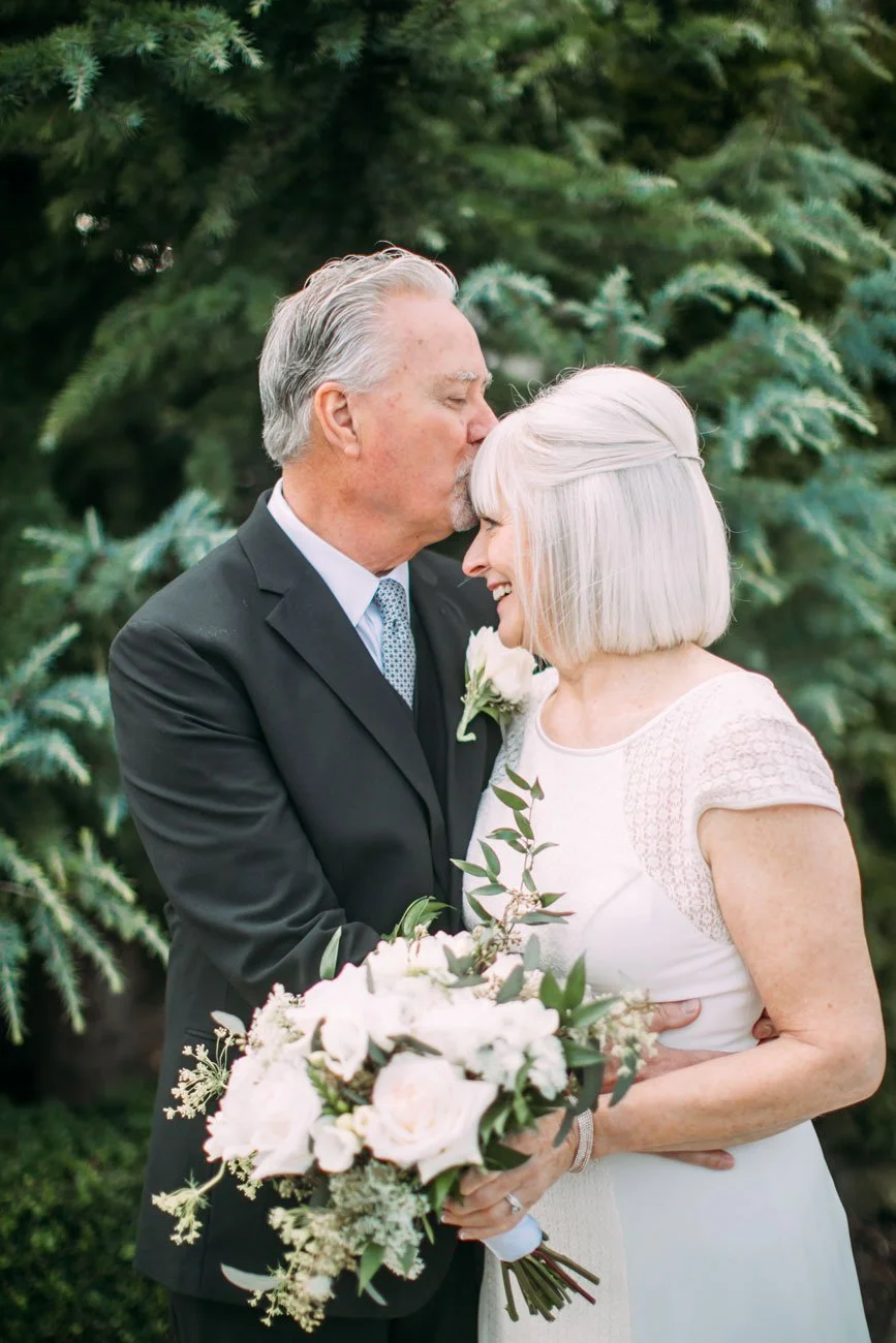 An elderly man in a black suit kisses an elderly woman with shoulder-length white hair on her forehead. The woman is smiling and holding a bouquet of white flowers with greenery. They are outdoors in front of green pine trees, sharing a tender moment