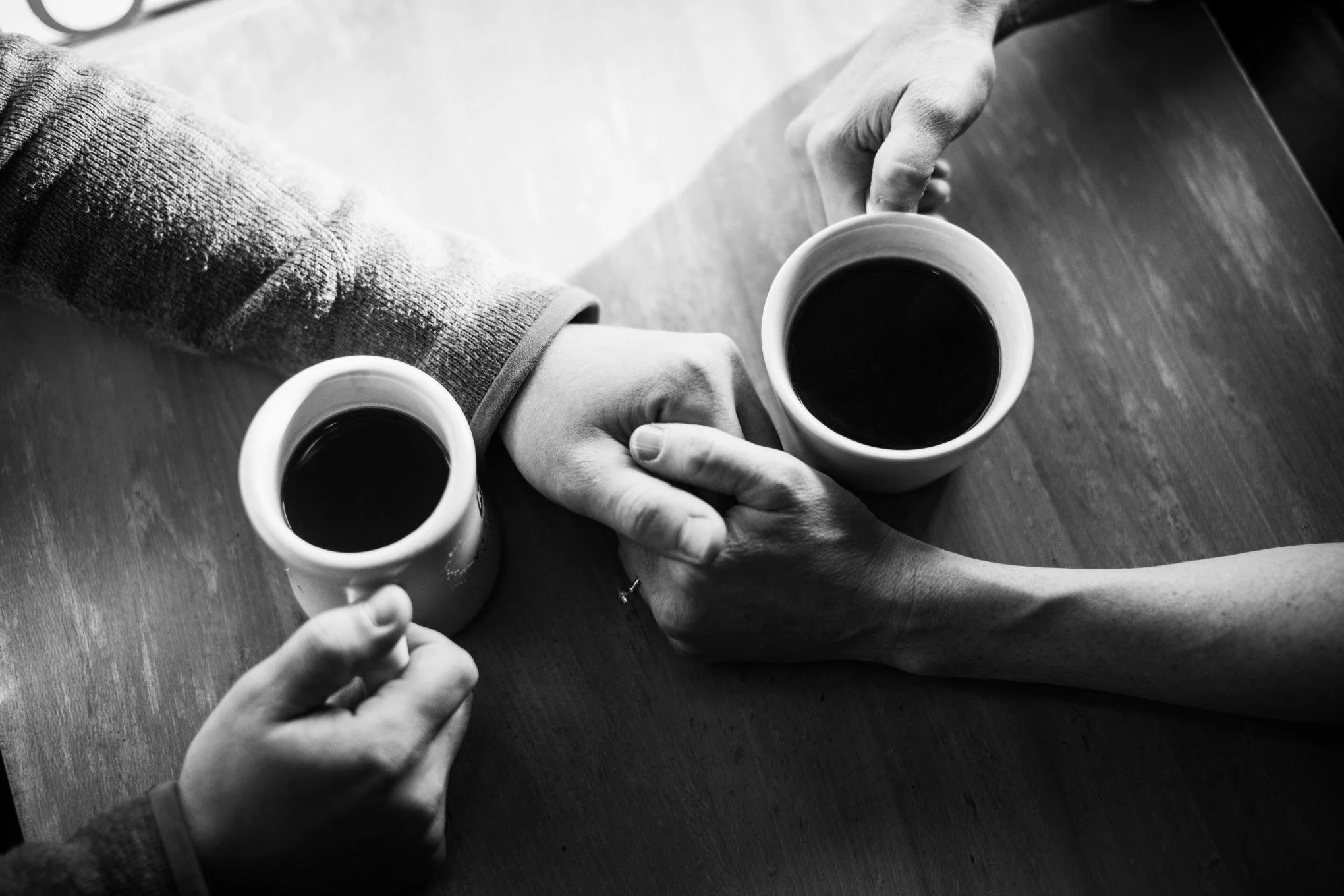 Two people holding matching mugs filled with dark coffee, clinking them together over a wooden table, in black and white.