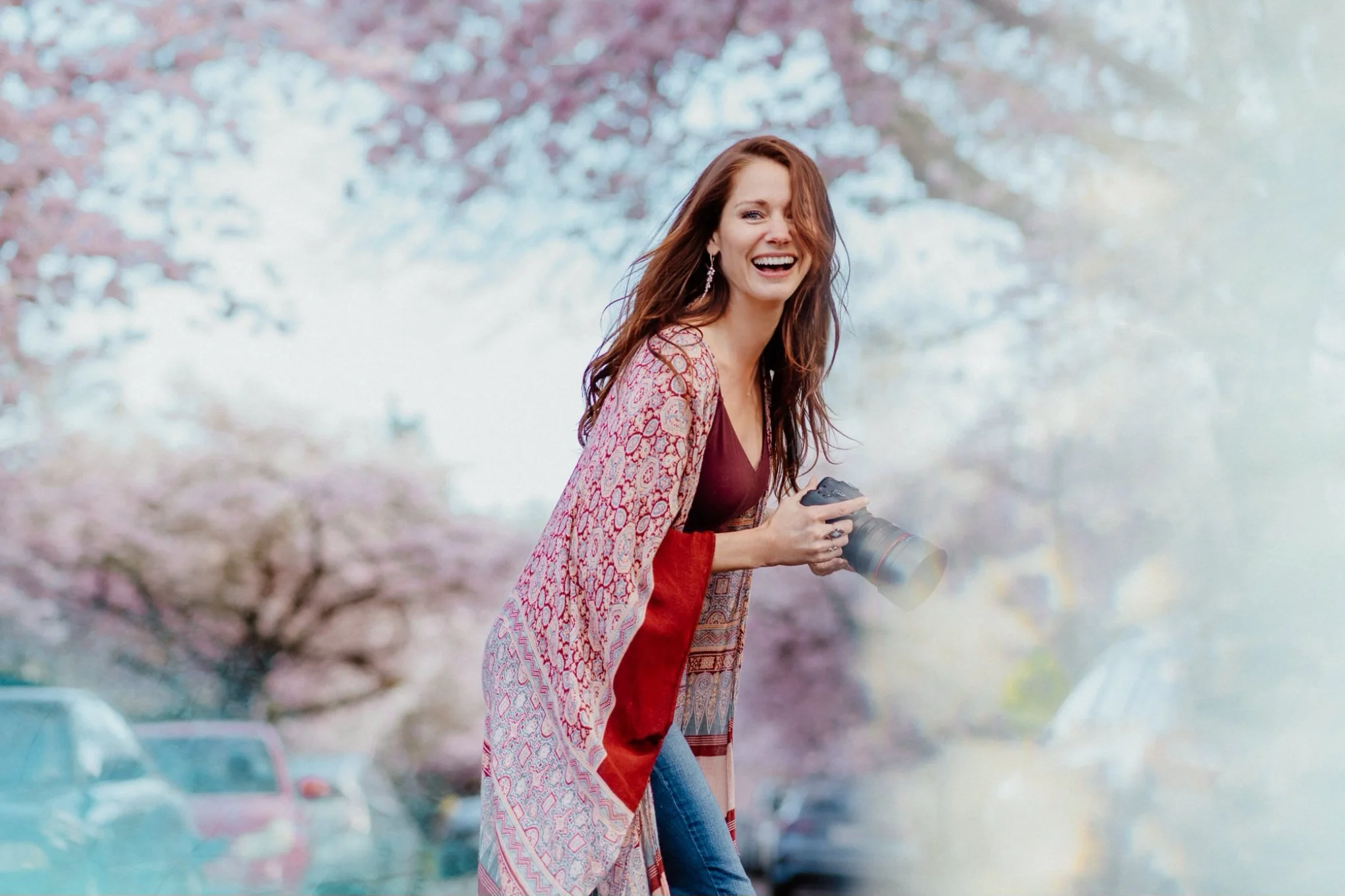 A woman with long, wavy auburn hair smiling and holding a camera, standing outdoors with pink cherry blossom trees and parked cars in the background in Seattle, WA.