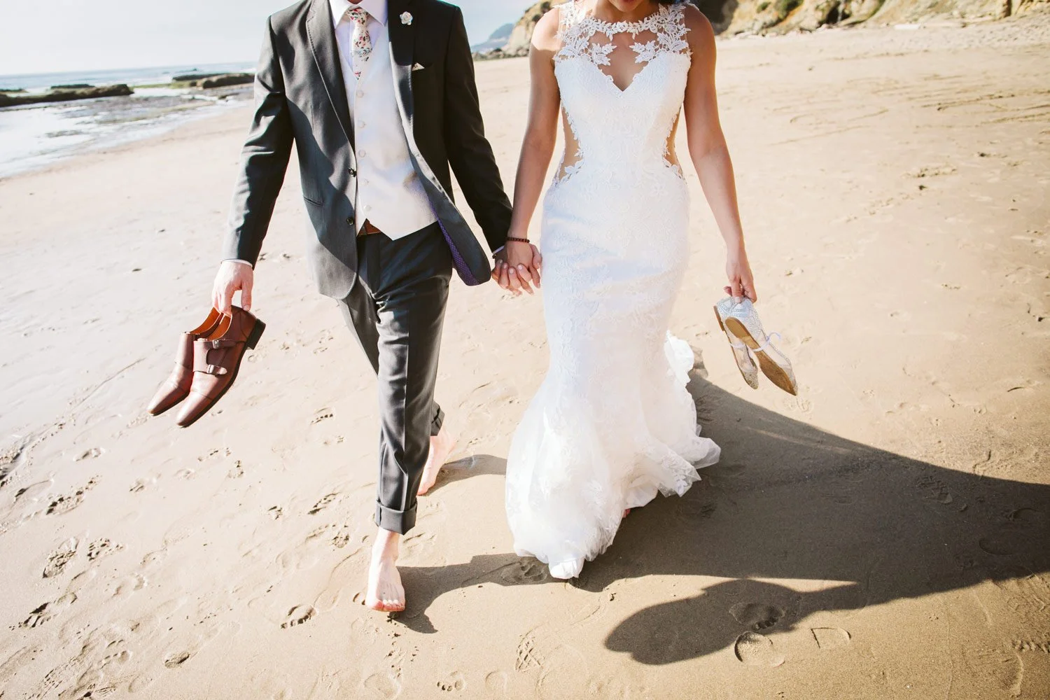 A newlywed couple holding hands walking barefoot on a sandy beach in San Diego, with the groom holding shoes in one hand and the bride carrying her shoes in the other, with the ocean and California coastline in the background.
