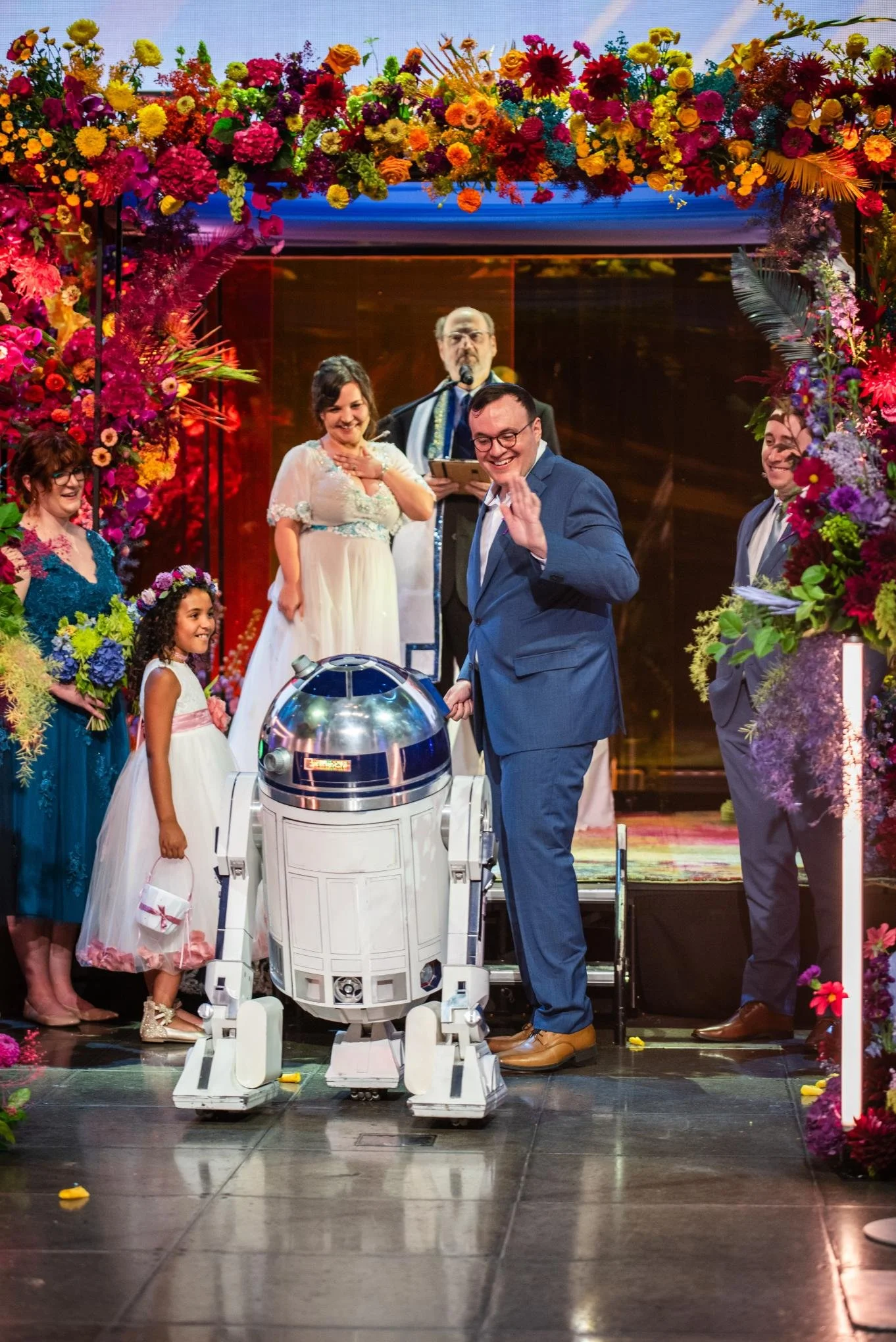 People in formal attire at a wedding ceremony over a floral arch, with a robot in the center and a man waving.