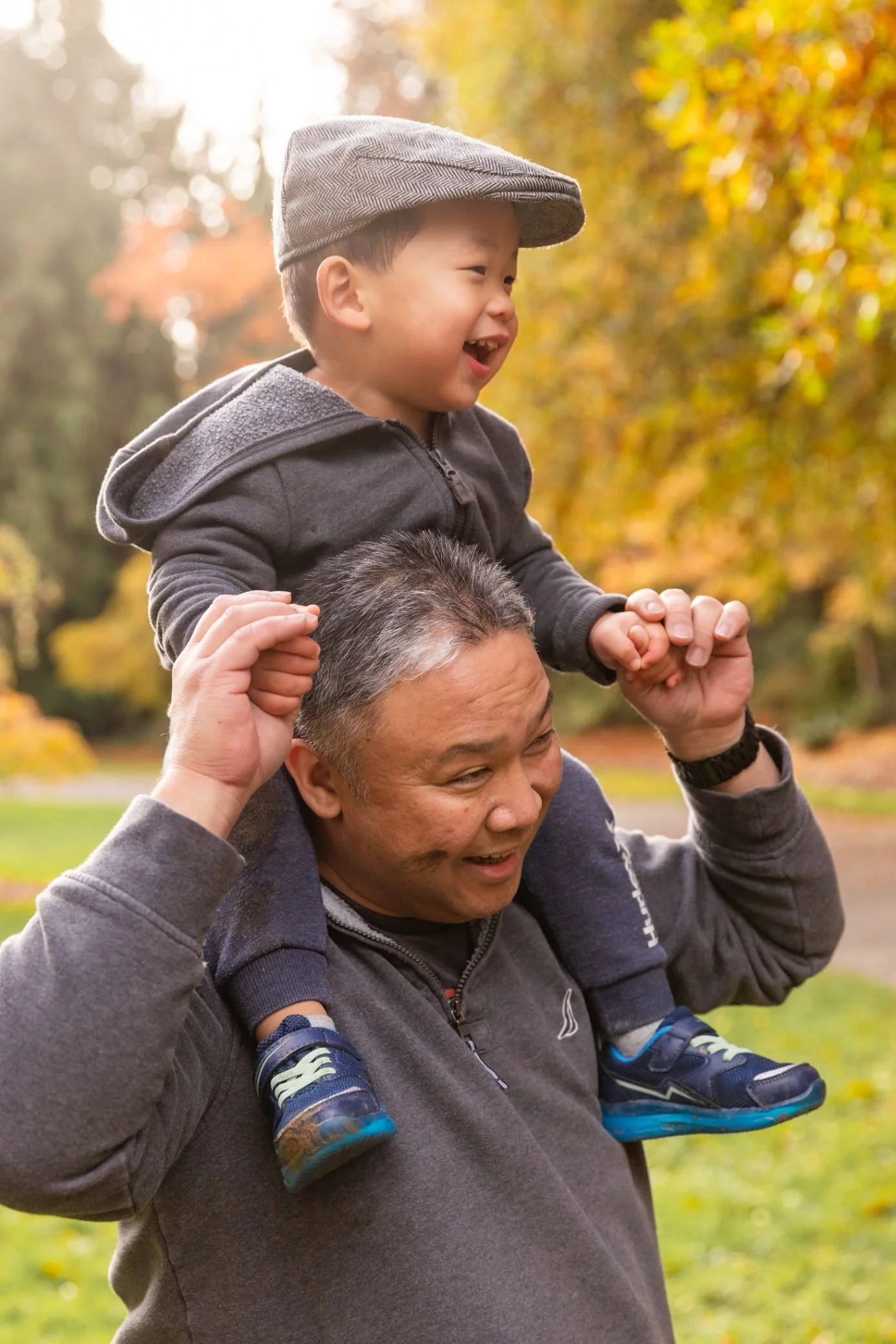 A man carrying a boy on his shoulders outdoors in a park with autumn trees in the background.