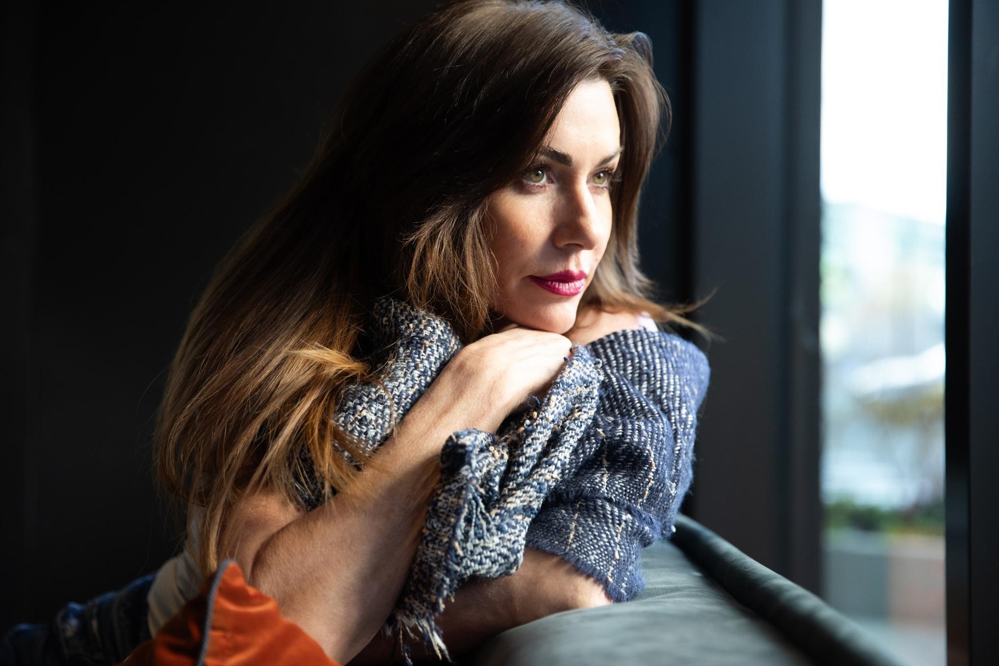 A headshot of a woman with brown hair and green eyes looking out of a window, resting her chin on her crossed arms, wearing a textured blue sweater.