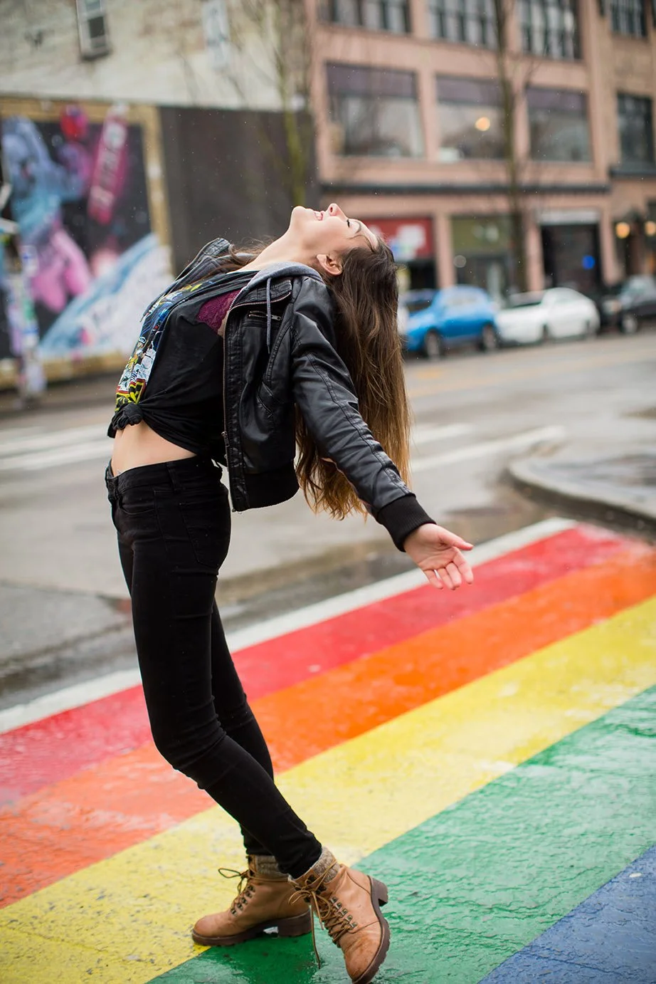 Person standing on a rainbow-colored crosswalk outdoors, with their head tilted back and arms extended, on a drizzly day.