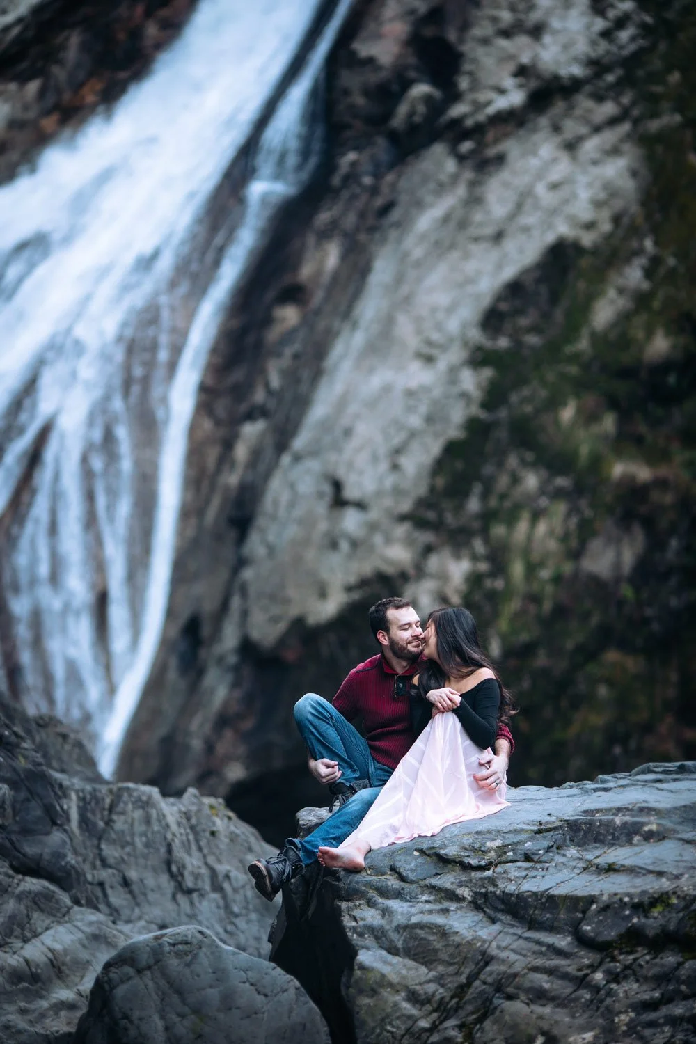 A couple sitting on rocks near a waterfall, sharing a kiss and embracing in a natural outdoor setting.