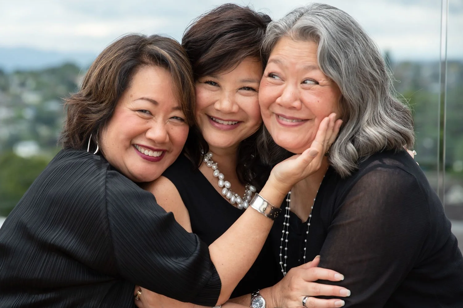 Three women happily hugging and smiling together outdoors with a cloudy sky and a blurred landscape in the background.