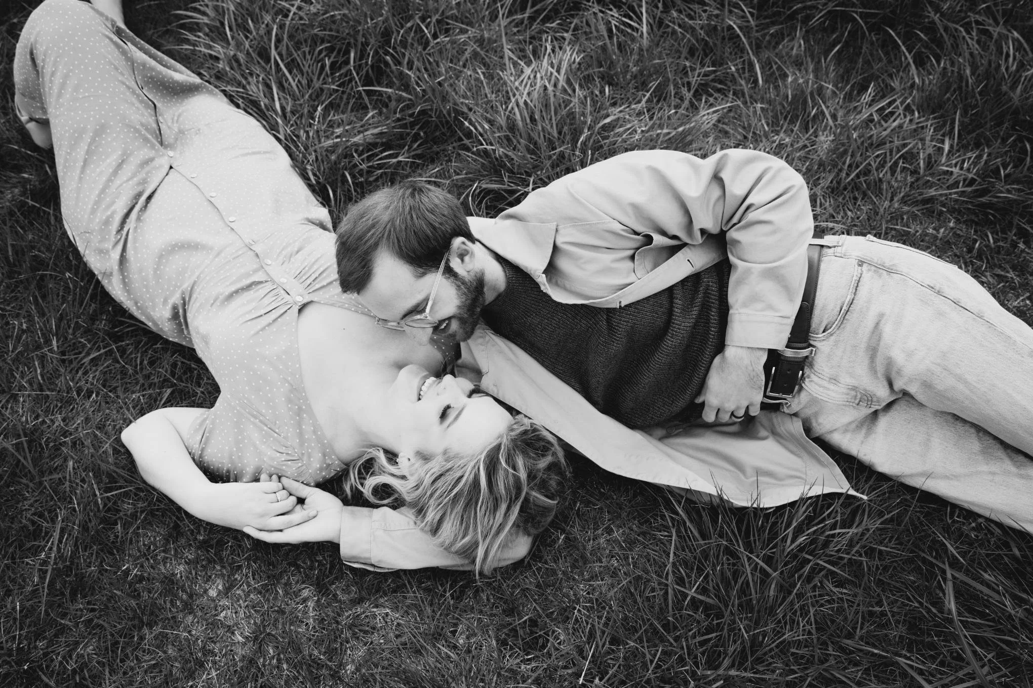 A black and white photograph of a young man and woman lying on grass, facing each other, smiling and holding hands.
