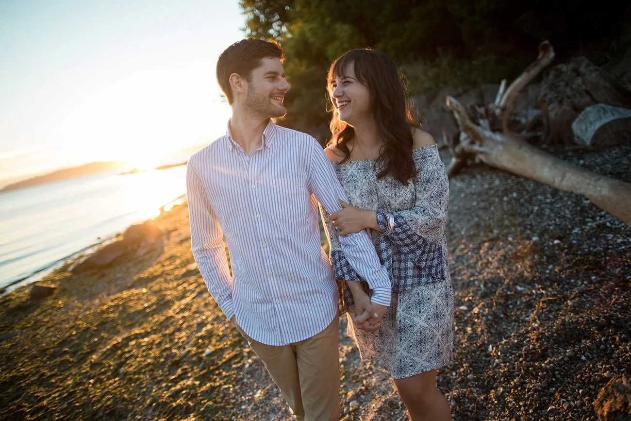 A smiling couple holding hands on a beach during sunset, with the sea, rocks, and driftwood in the background.