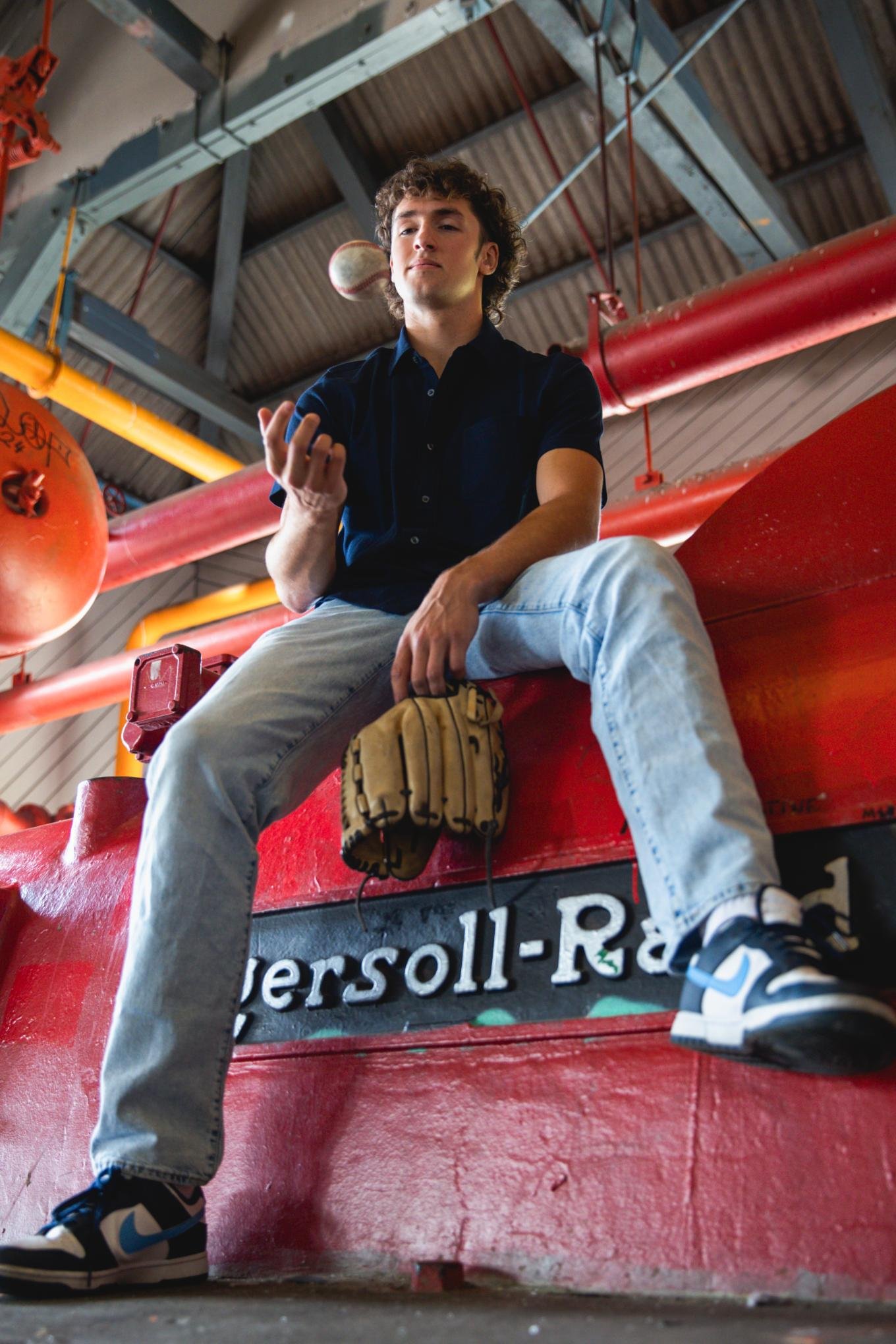 A young man with curly brown hair, wearing a navy blue shirt and light blue jeans, sits on a red industrial structure inside a fire station. He holds a baseball glove in his left hand and is looking at a baseball or softball in his right hand, which 