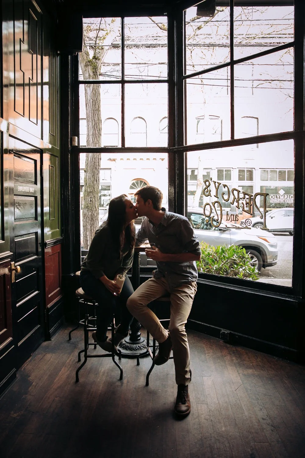 A couple sharing a kiss inside a cafe, sitting on stools near a large window with street view, trees, and parked cars outside.