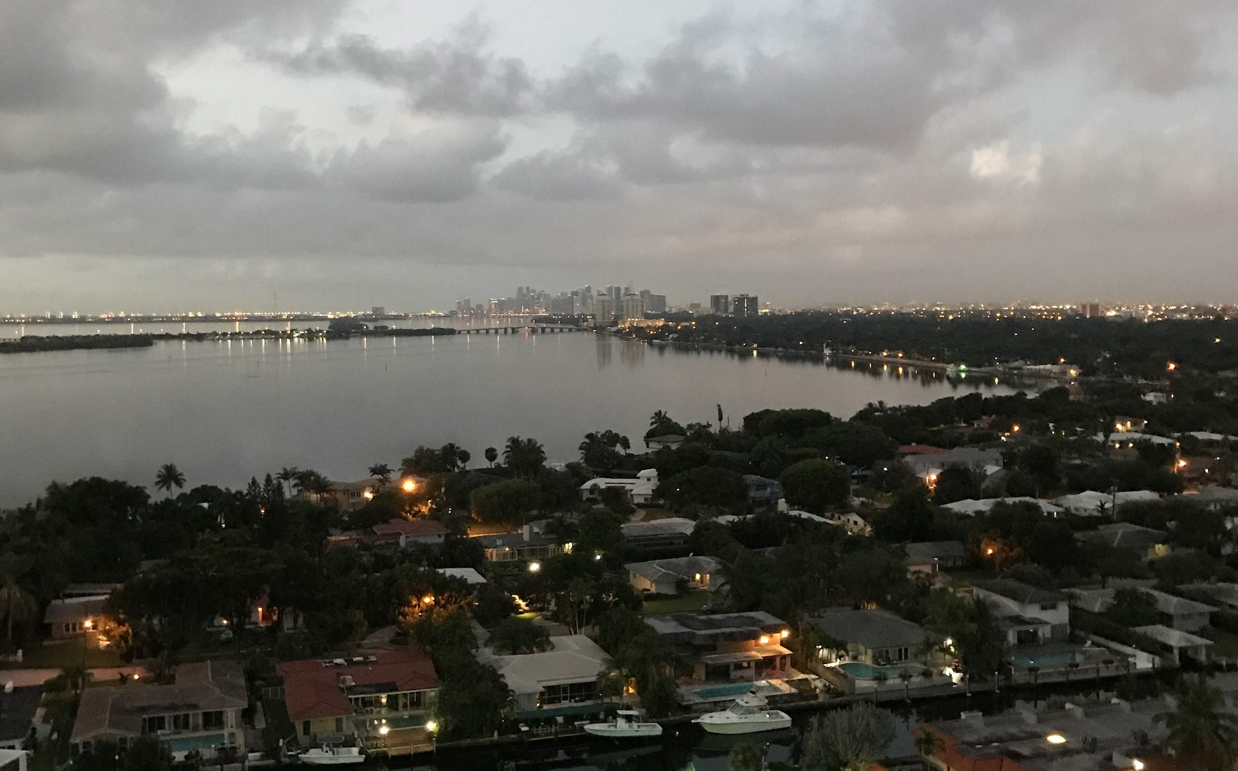 Miami Shores Aerial view of a coastal cityscape at dusk, featuring a large body of water with surrounding residential areas. In the background, a skyline with high-rise buildings is visible under a cloudy sky.