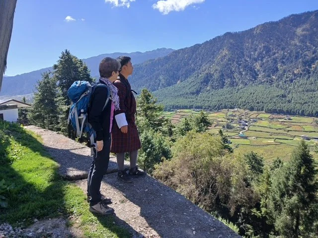 A traveler and Bhutanese guide explore Bhutan’s scenic countryside, admiring lush landscapes, traditional villages, and cultural sites. Experience guided walking tours, trekking, and local insights.