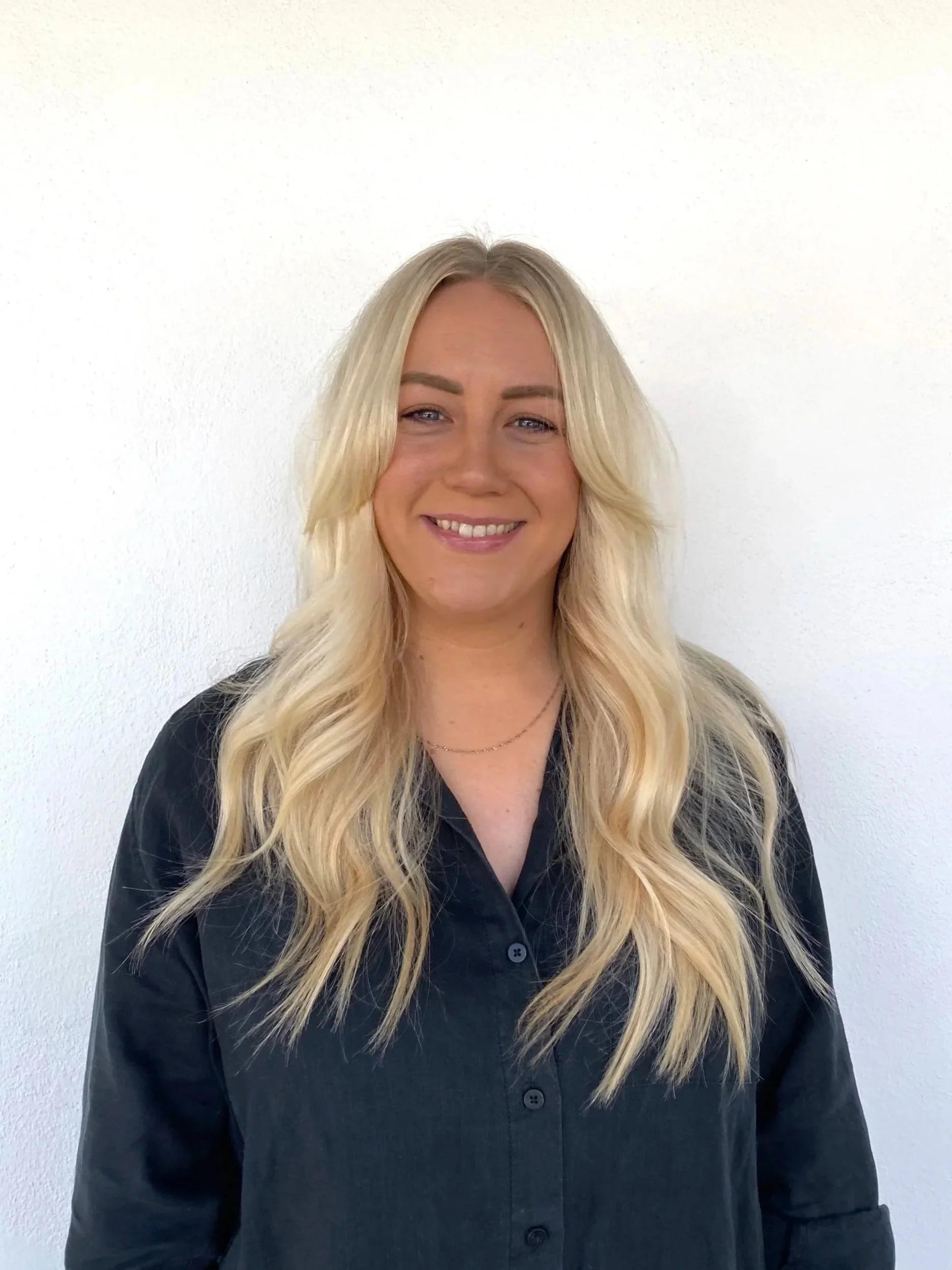 A woman with long wavy blonde hair, wearing a black button-up shirt, smiling in front of a white wall.