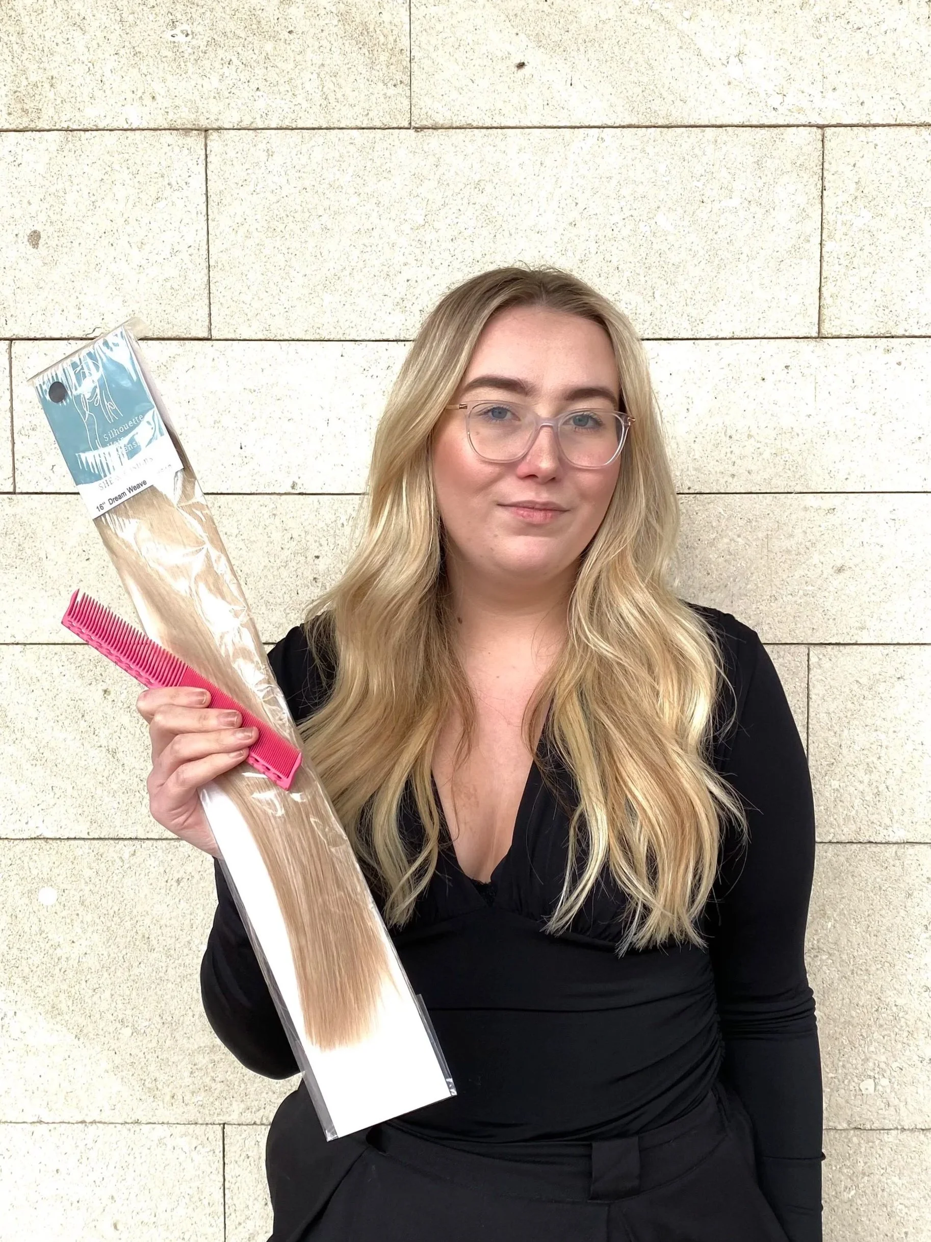 A woman with long blonde hair, wearing glasses and a black top, holding a package of blonde hair extensions and standing in front of a beige brick wall.
