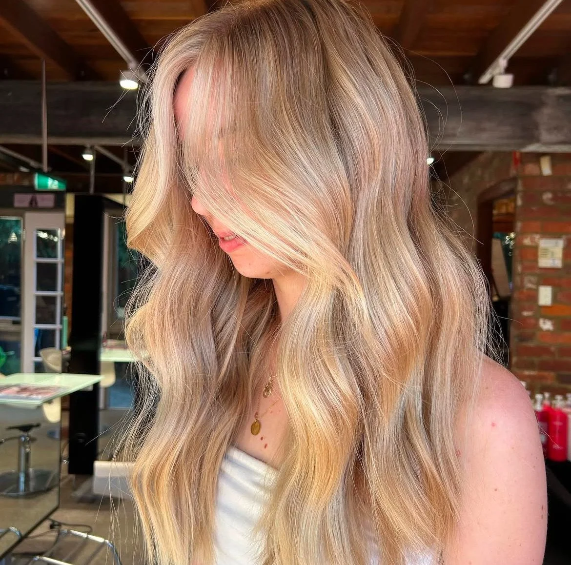 A woman with long, wavy blonde hair, with her face partially obscured by her hair, standing inside a salon with brick walls and wooden beams.