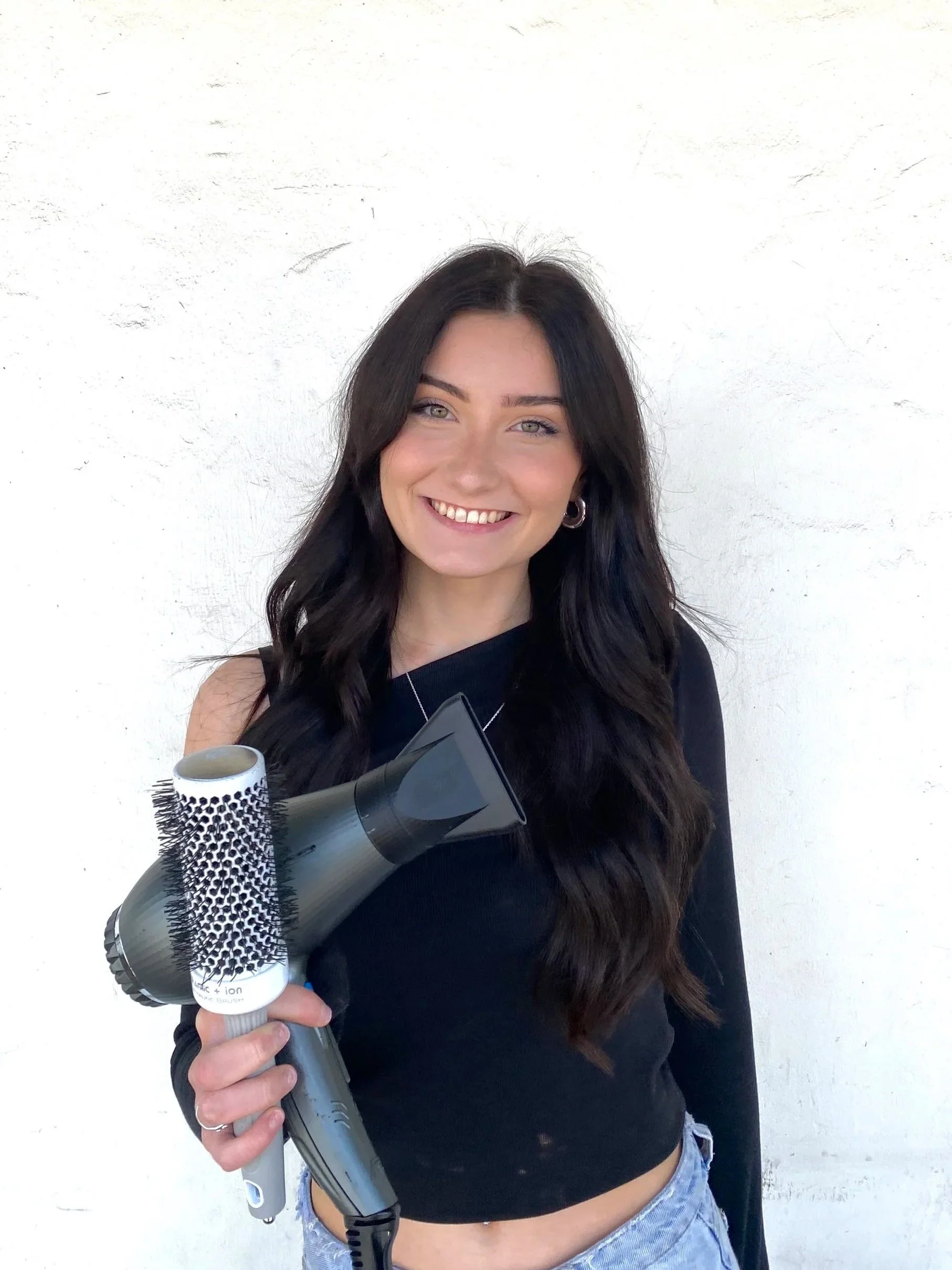 A young woman with long, dark wavy hair, smiling, holding a hairdryer and a round brush, standing against a white wall.