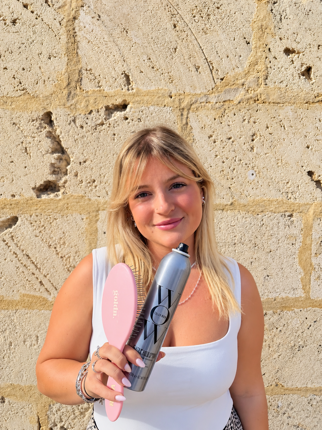 A young woman with long blonde hair holding a pink comb and a small purple gift box, standing against a beige stone wall.