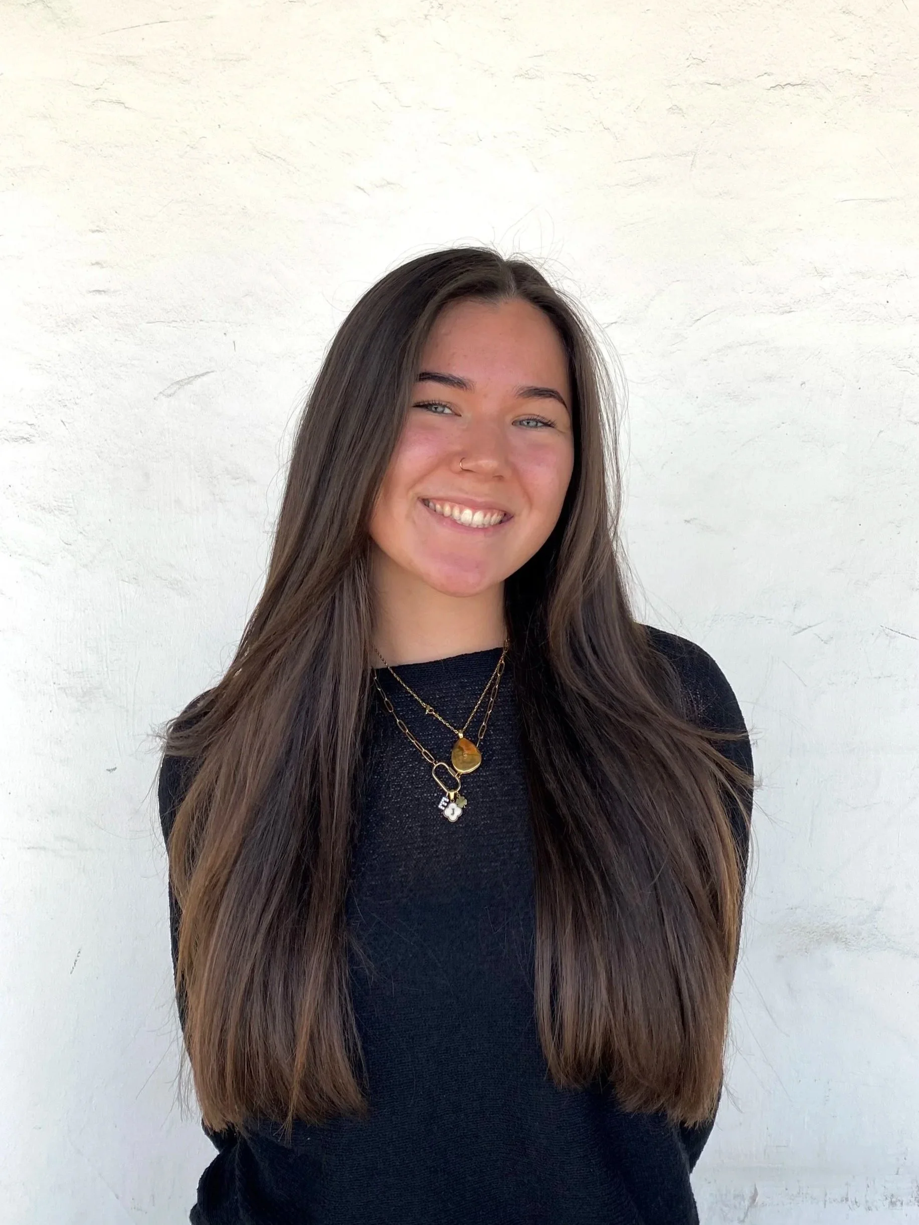 Young woman with long brown hair smiling, wearing layered necklaces, black top, standing against a plain white wall.