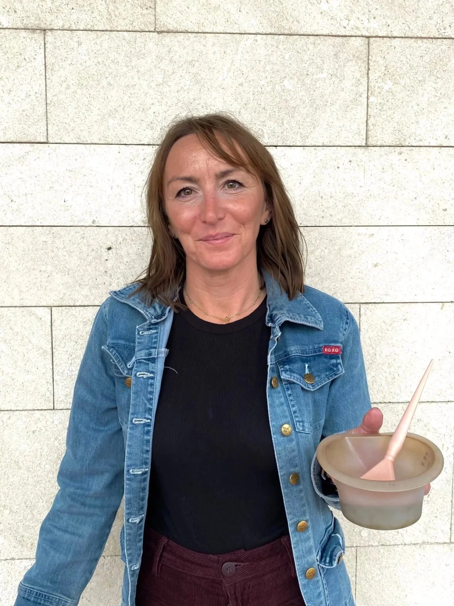 A woman with shoulder-length brown hair, wearing a denim jacket over a black shirt, holding a beige bowl with a pink spoon, standing against a white stone wall.