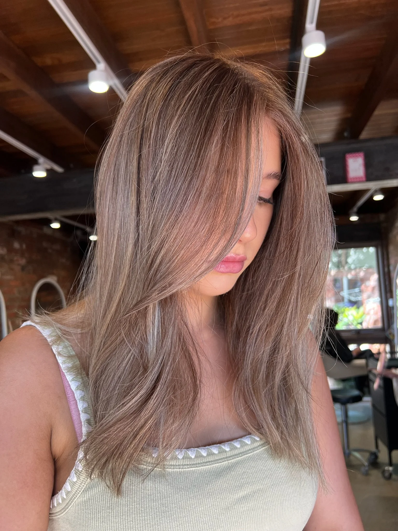 A woman with long, layered blonde hair with subtle highlights, looking downward in a brightly lit salon with wooden ceilings and brick walls.