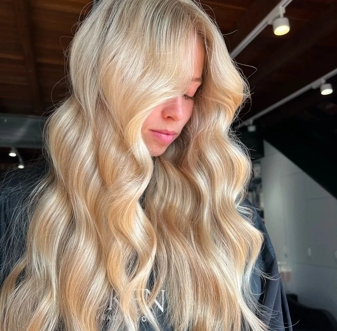 Close-up of a woman with long, wavy blonde hair looking down indoors with a wood ceiling and lights in the background.