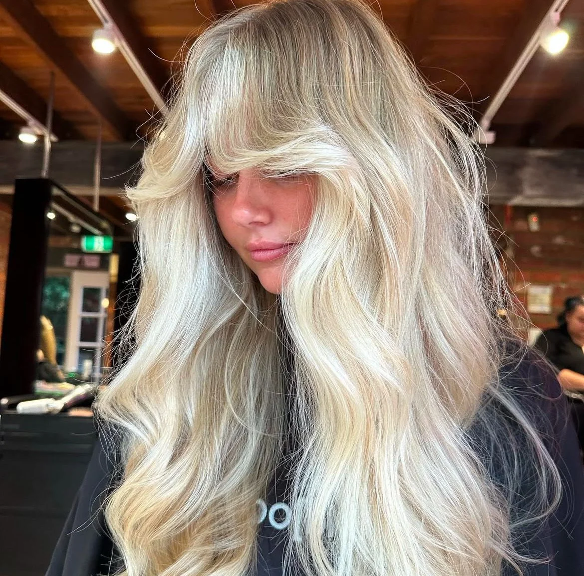 Woman with long, voluminous, wavy blonde hair in a salon or barbershop setting.