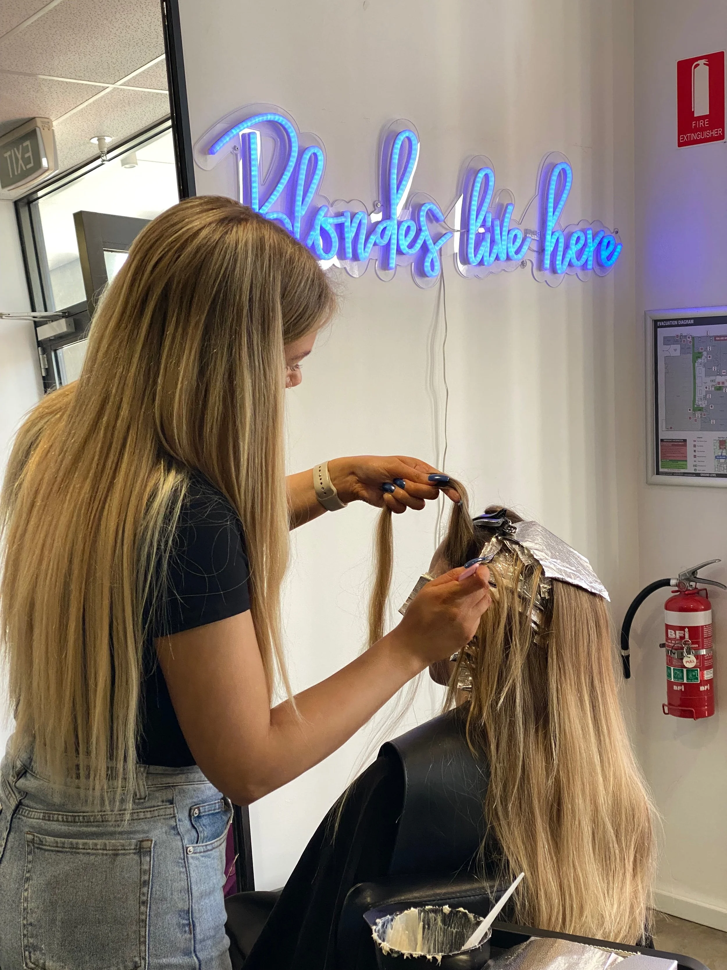 A woman with long blonde hair is applying hair dye to another woman's hair at a salon. The person receiving the treatment has foil in her hair. There is a neon sign on the wall that says 'Blondes live here' in blue cursive text. A fire extinguisher and a fire safety sign are visible on the wall.