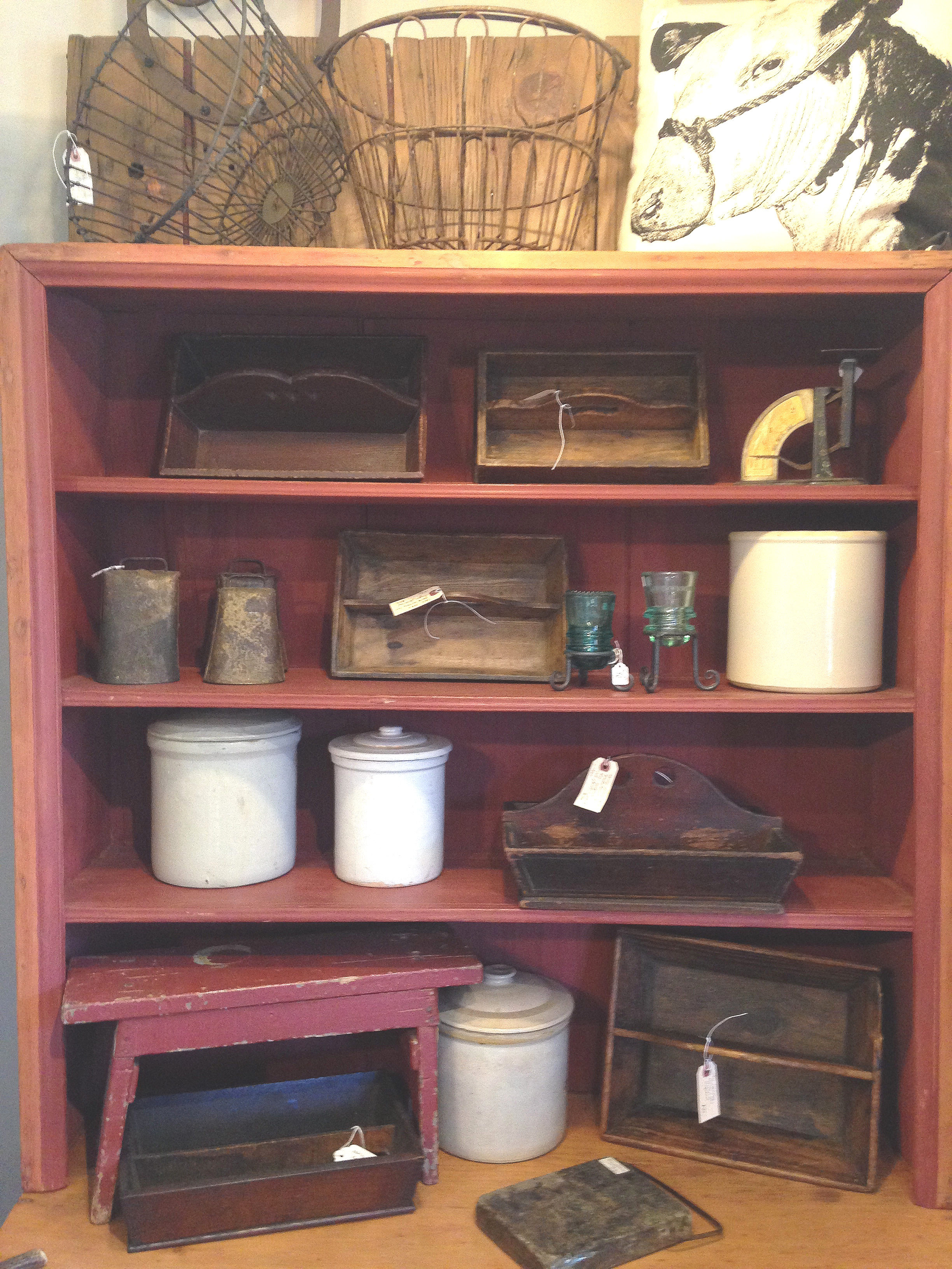 An assortment of antique knife/cutlery trays, stoneware crocks, cow bells, egg graders, and wire baskets round out this classic country display.