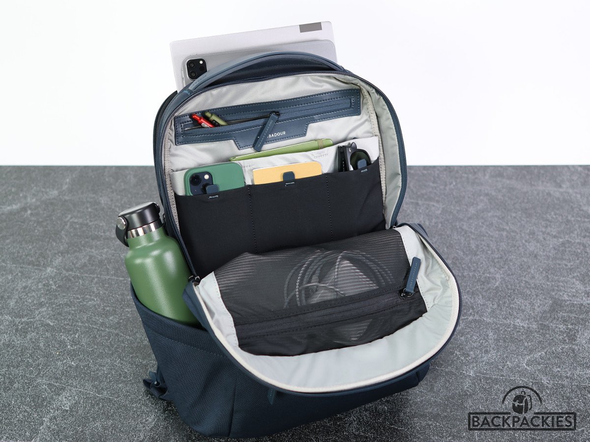 Photo of the open 25L main compartment of the Troubadour Apex men's backpack for work, revealing internal pockets filled with a small notebook, pens, a hard drive, a phone, and a laptop charger. The external side pocket holds a water bottle.