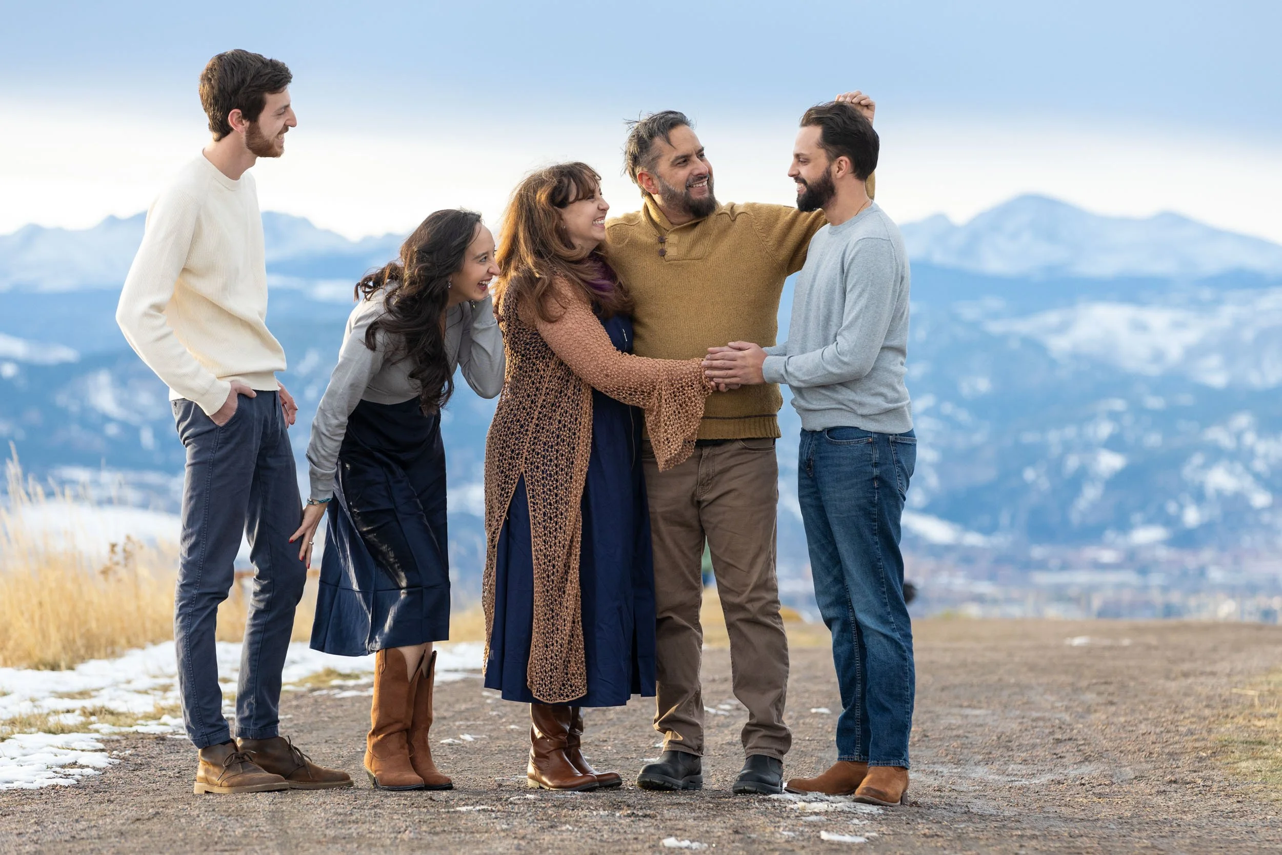 A group of six people standing outdoors on a mountain trail, enjoying each other's company with mountains in the background. They are smiling, laughing, and appear to be sharing a happy moment.
