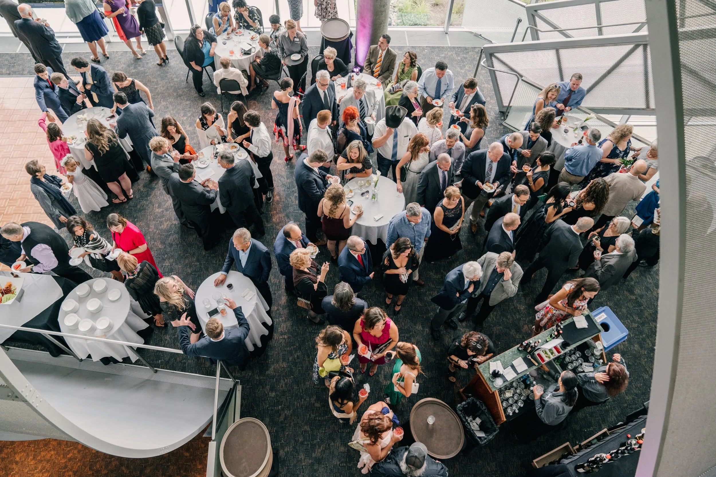 A large crowd of people attending a social event or reception in an indoor venue. The attendees are mingling around tables, some standing in line at a bar, and others eating or drinking.