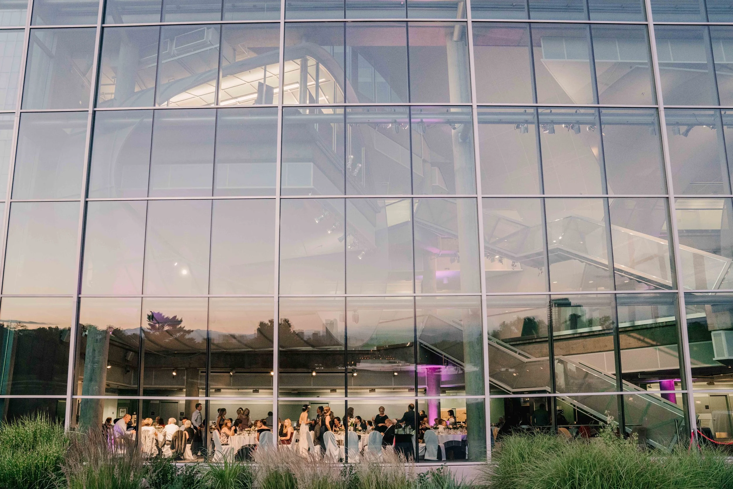 An event taking place inside a modern glass building with large windows, showcasing multiple round tables with white tablecloths, people seated and chatting, and some standing waitstaff. The building has a staircase visible through the glass, with pu