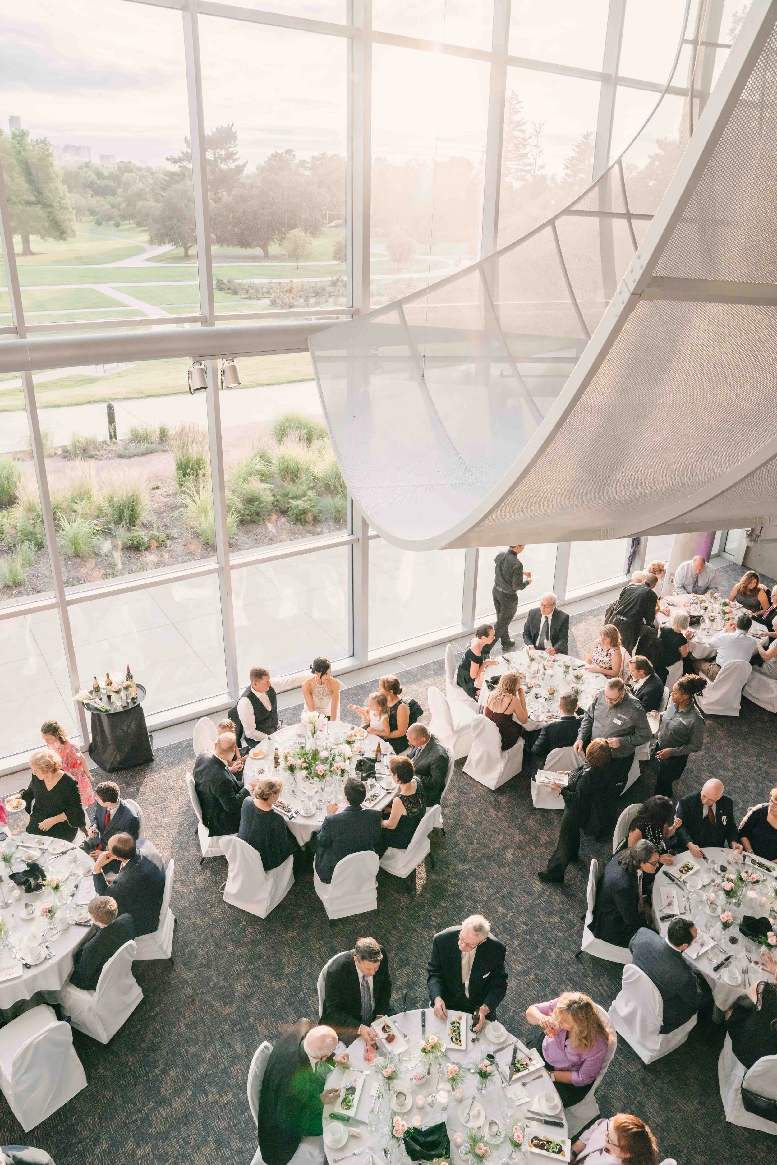 A large, brightly lit banquet hall filled with round tables with white tablecloths and floral centerpieces, guests dressed in formal attire, and floor-to-ceiling windows overlooking a park with trees and pathways.