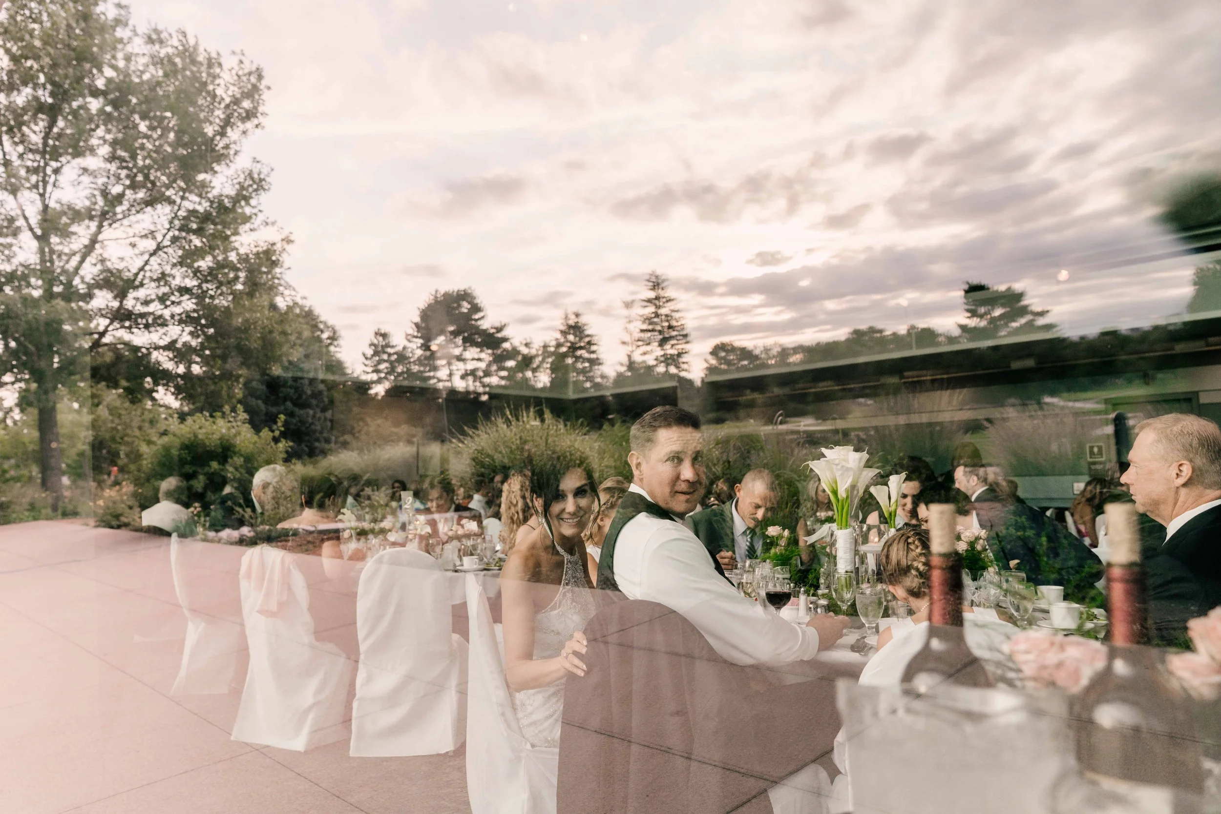 A glass window reflecting a wedding reception outdoor where guests are seated at a long table with white chairs, floral centerpieces, and wine bottles. The outdoor setting includes trees and a partly cloudy sky.