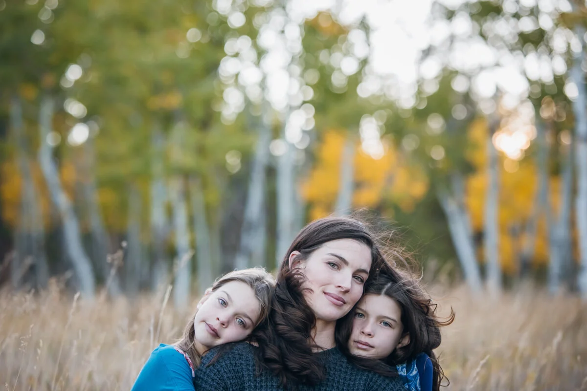 A woman with two young girls in a field with trees and autumn leaves in the background.