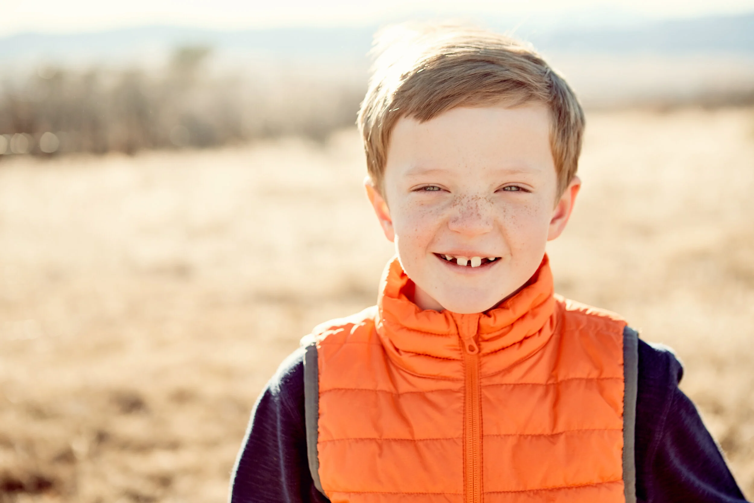 Smiling child in orange vest outdoors with sunny background.