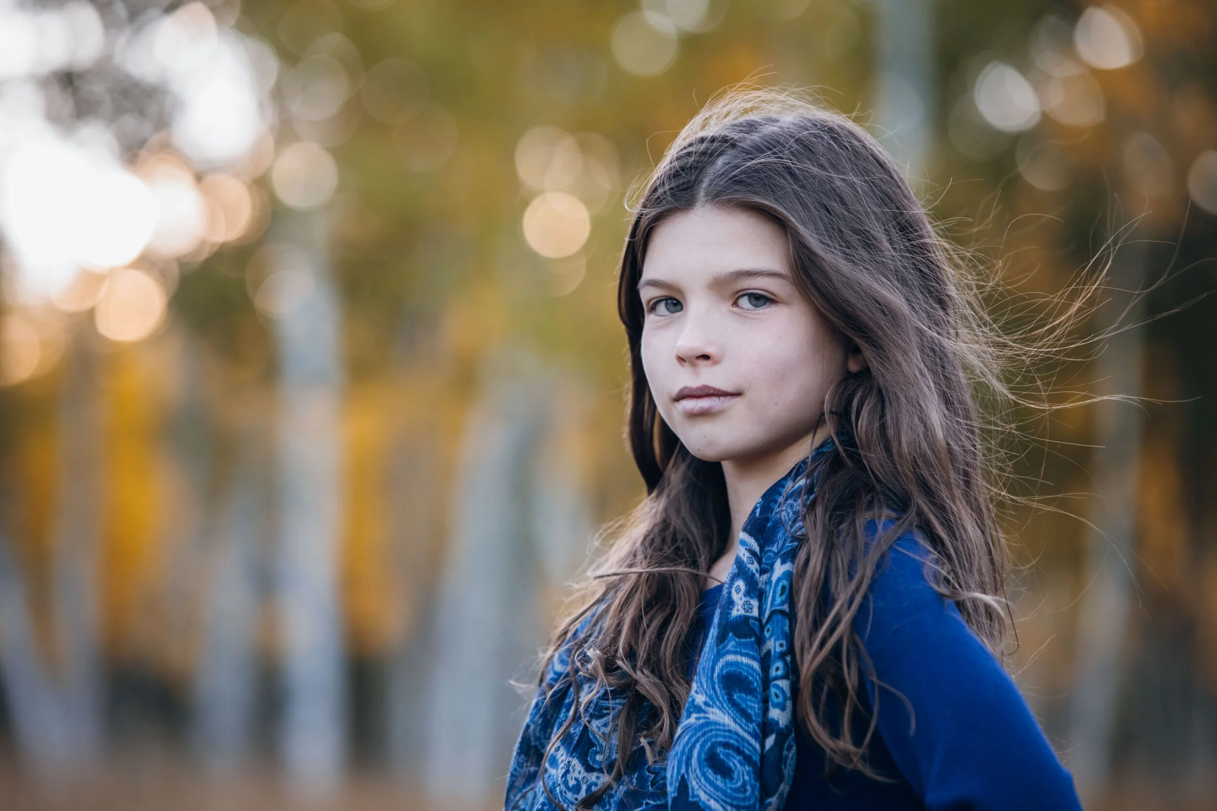 Girl with long brown hair wearing blue scarf in autumn forest.