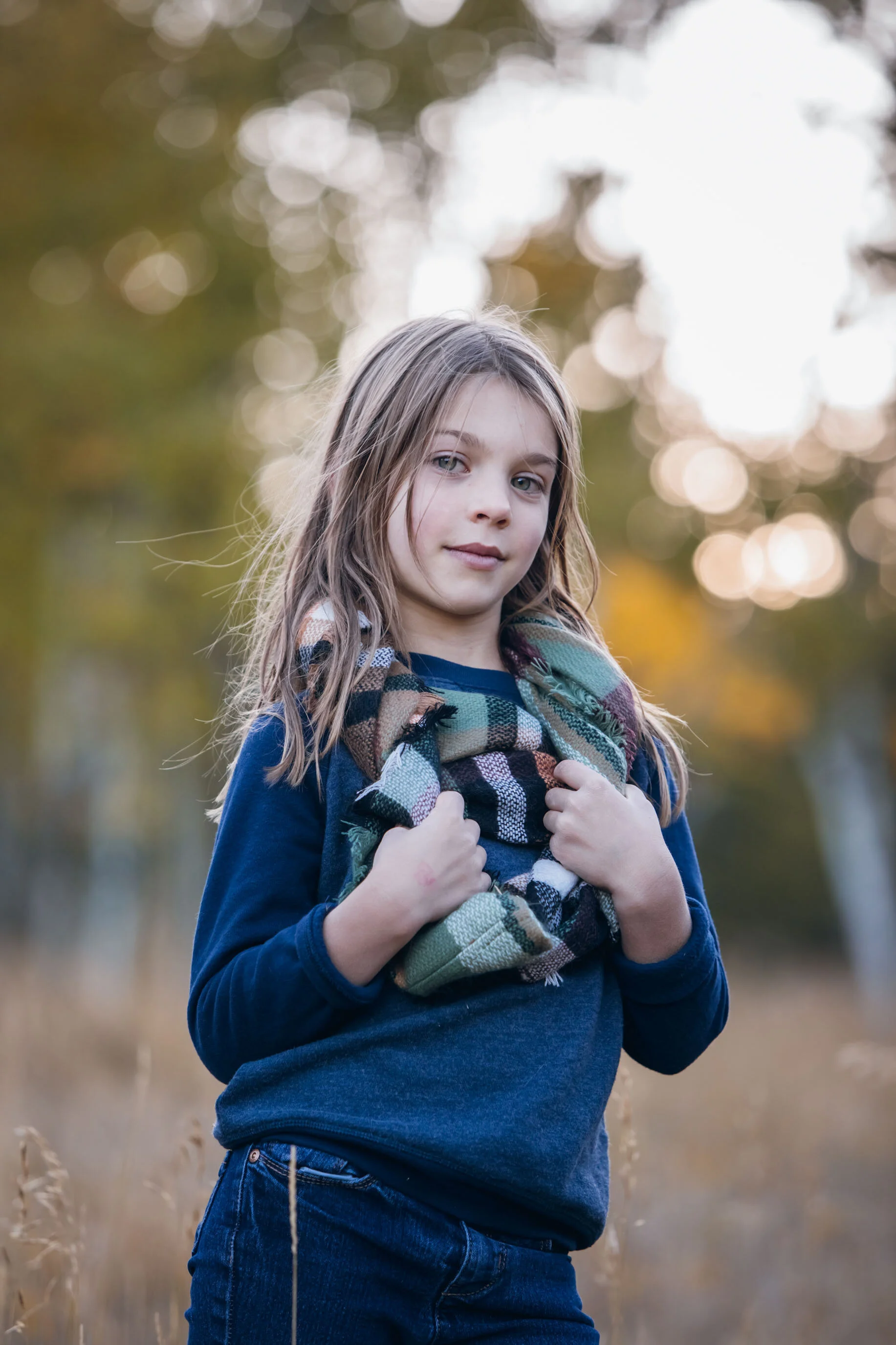A young girl with long hair wearing a blue sweater and checkered scarf stands outdoors with a blurred autumn background.