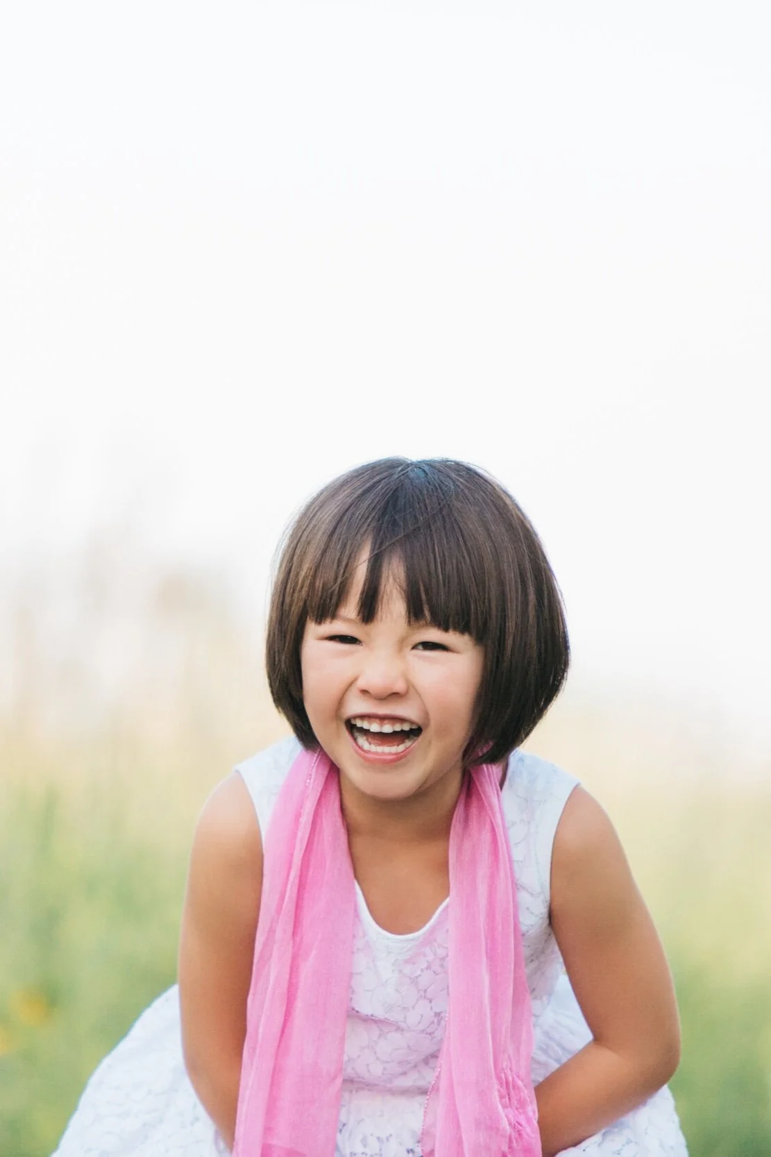 Young girl laughing, wearing a pink scarf and white lace dress, outdoors.