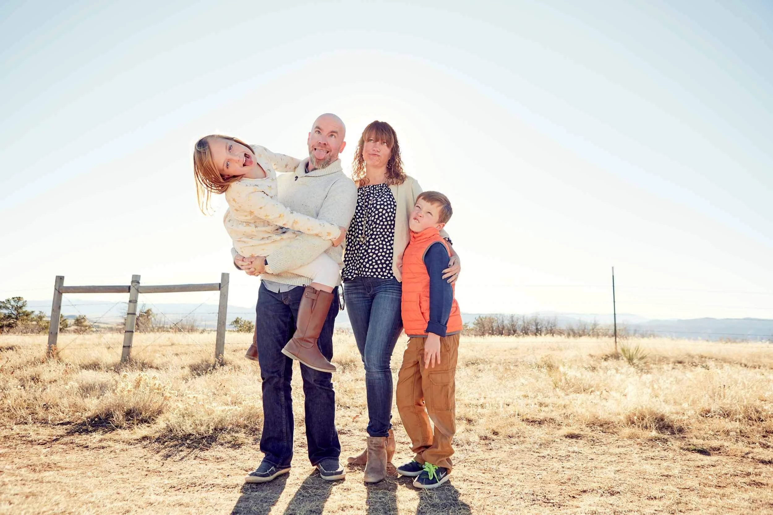 A family of four standing outdoors in a dry field with hills in the background, with sunlight coming from behind them.
