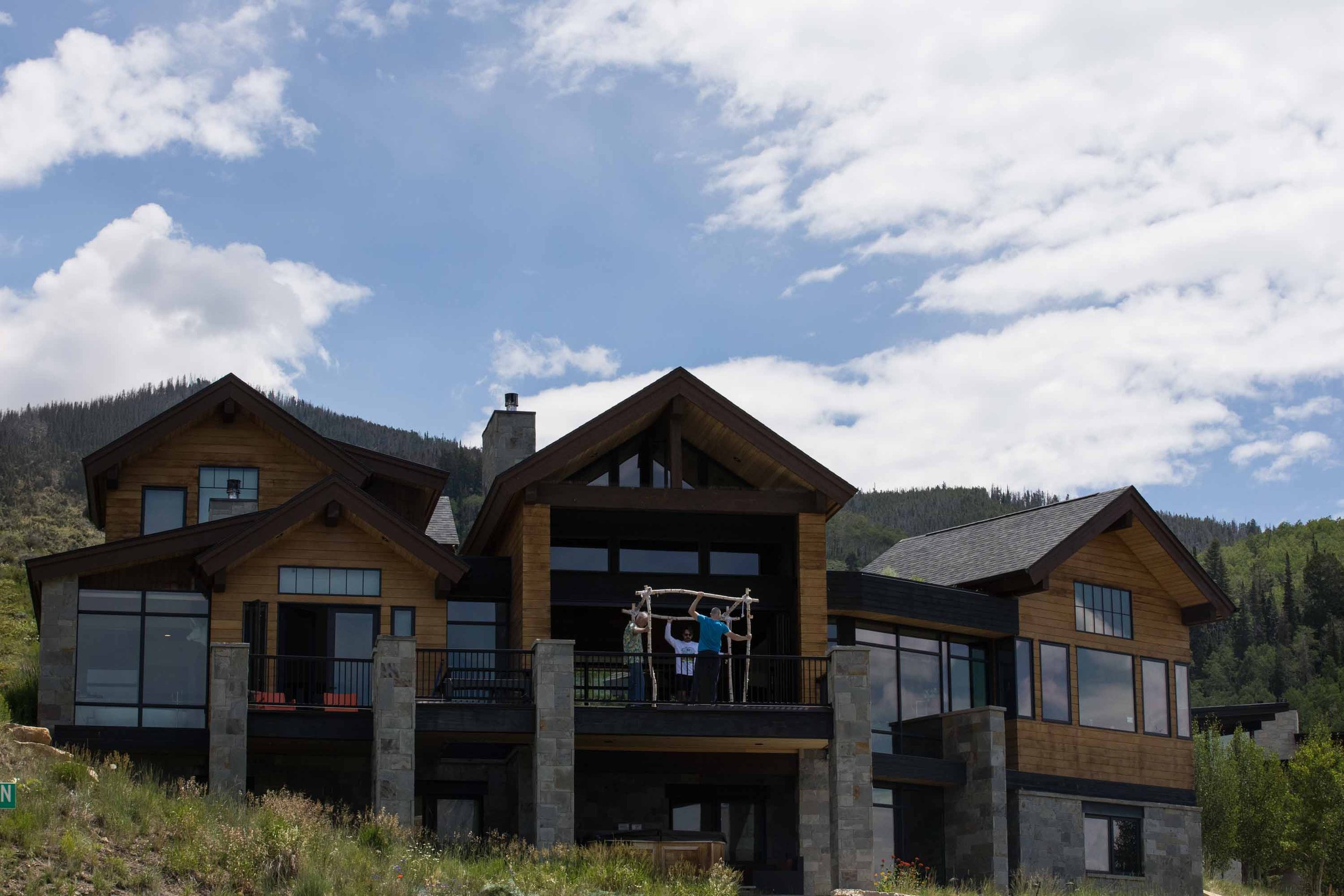 A large modern house with wooden and stone exterior, multiple large windows, and a balcony. Two children and an adult are on the balcony, hanging a large wooden frame or structure, with mountains and a partly cloudy sky in the background.