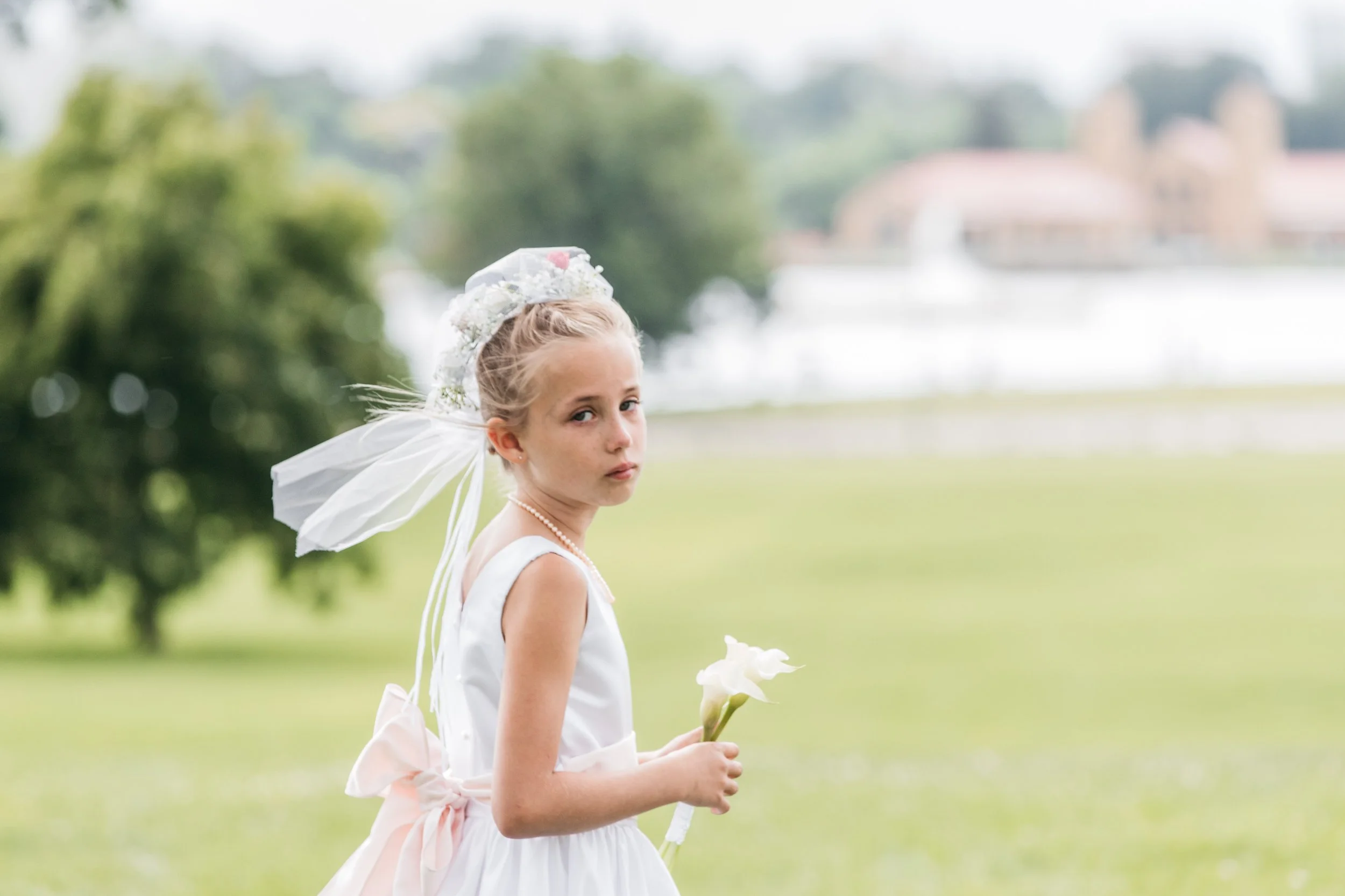 A young girl dressed in a white dress and with a floral headpiece, holding a white flower, standing outdoors on a grassy field with trees and buildings in the blurred background.